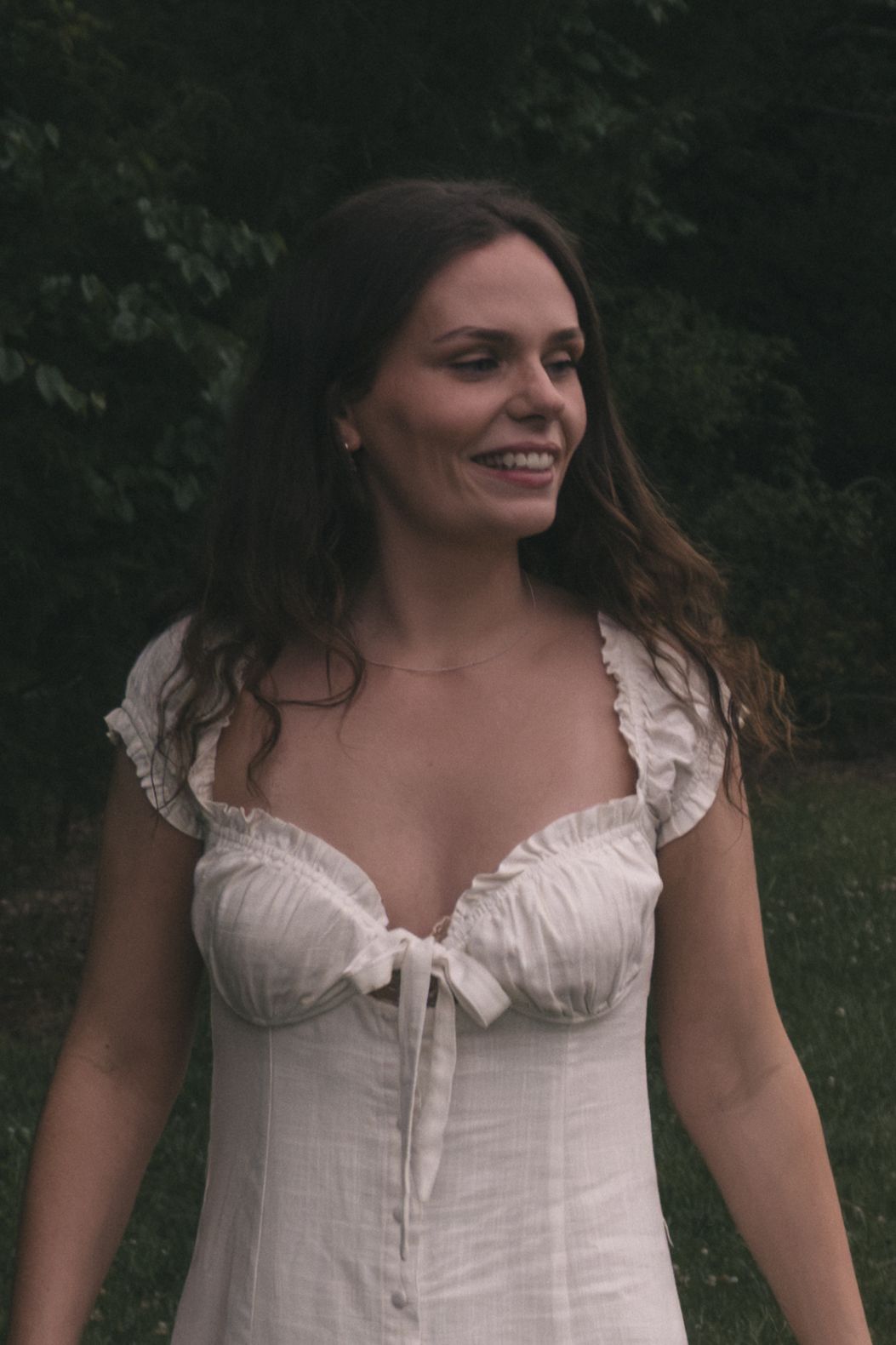 A smiling brunette woman in a white ruffled sundress stands outdoors with green trees in the background.