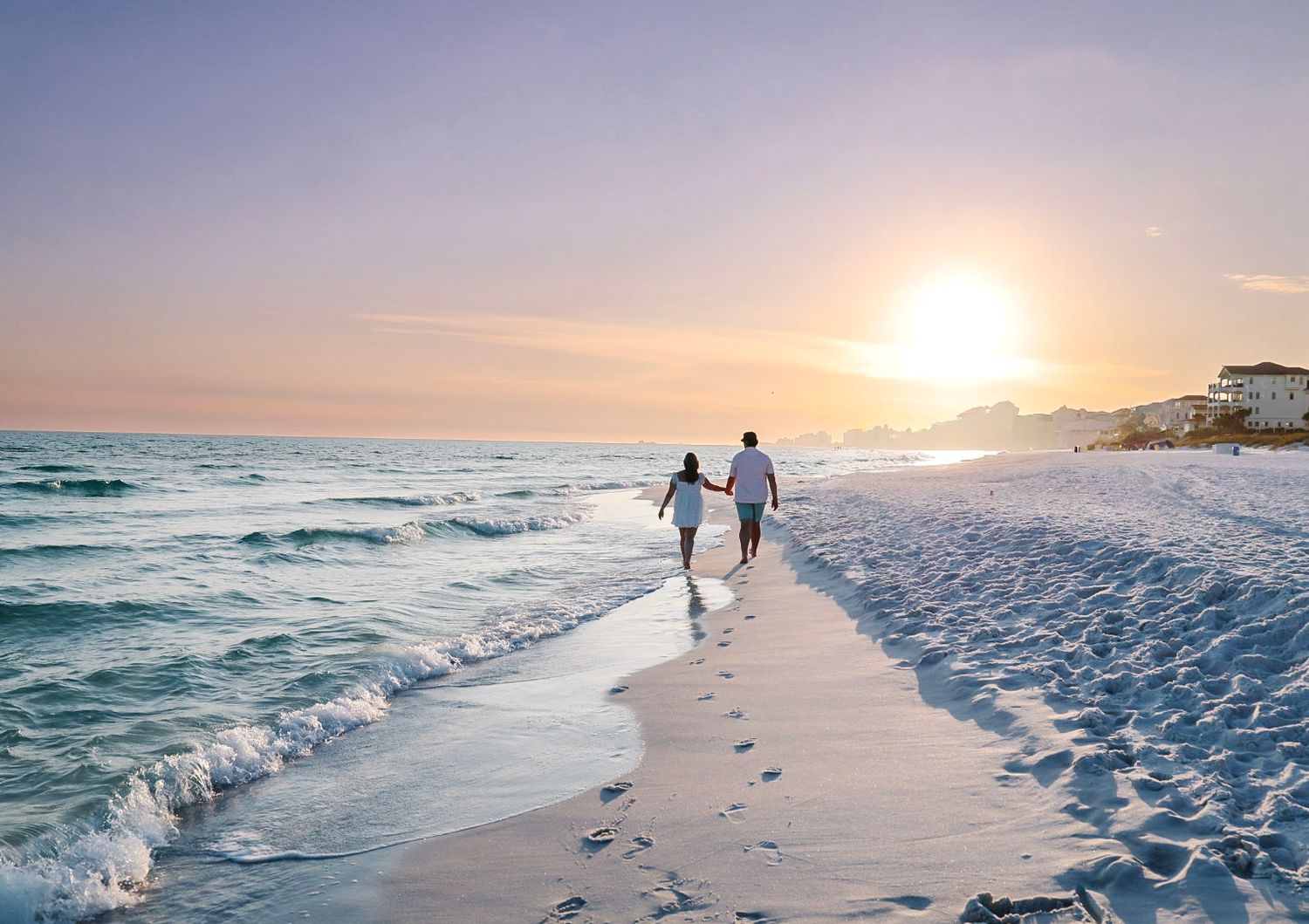 Couple holding hands walking along a sandy beach at sunset with gentle waves and pastel skies.
