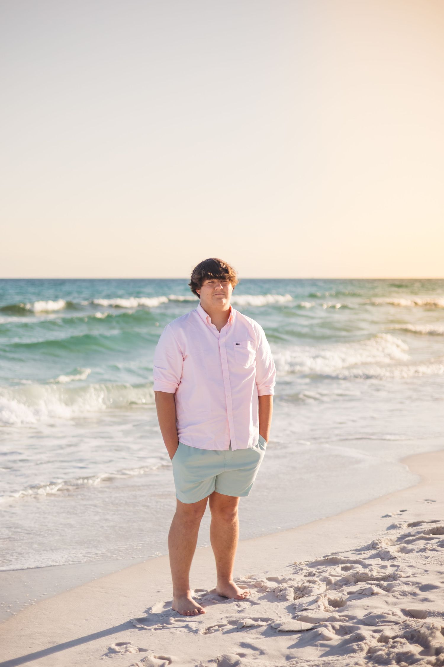 Young man in pink button-up and mint shorts poses on white sand beach at sunset.