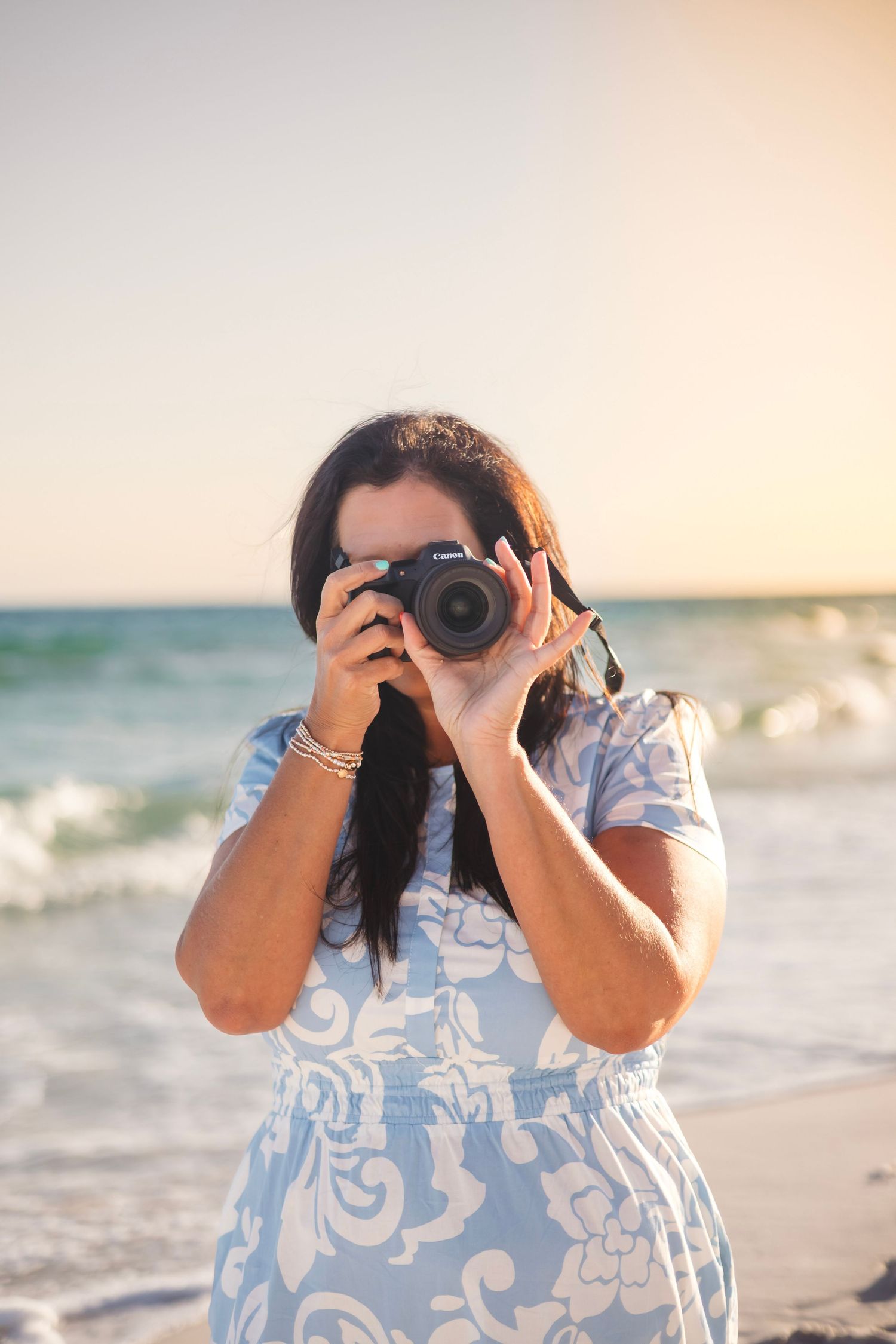 Woman in blue floral dress photographing with Canon camera on a sunny beach with ocean waves behind her.