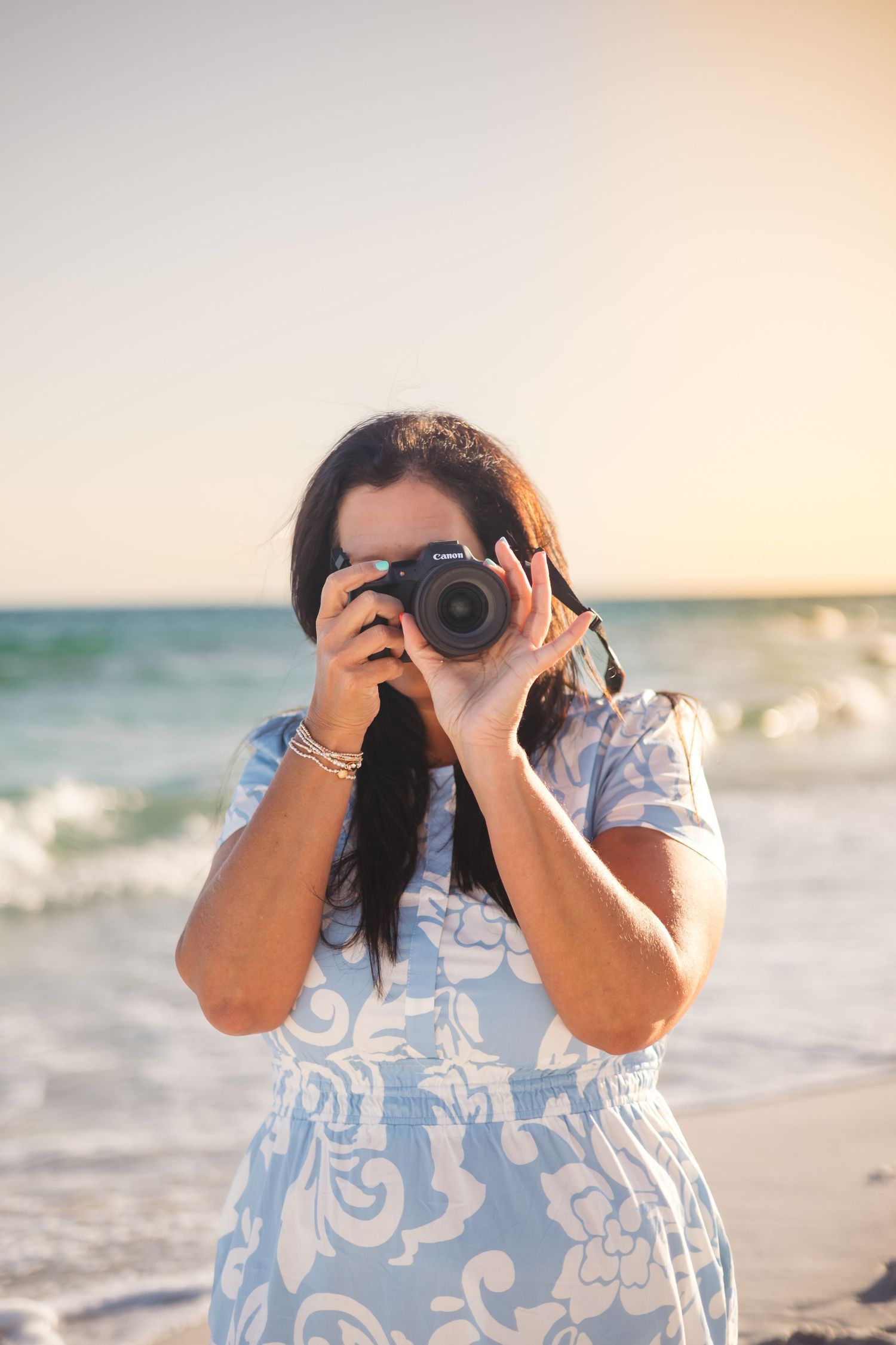 Woman in blue floral dress photographs with Canon camera on beach at golden hour.