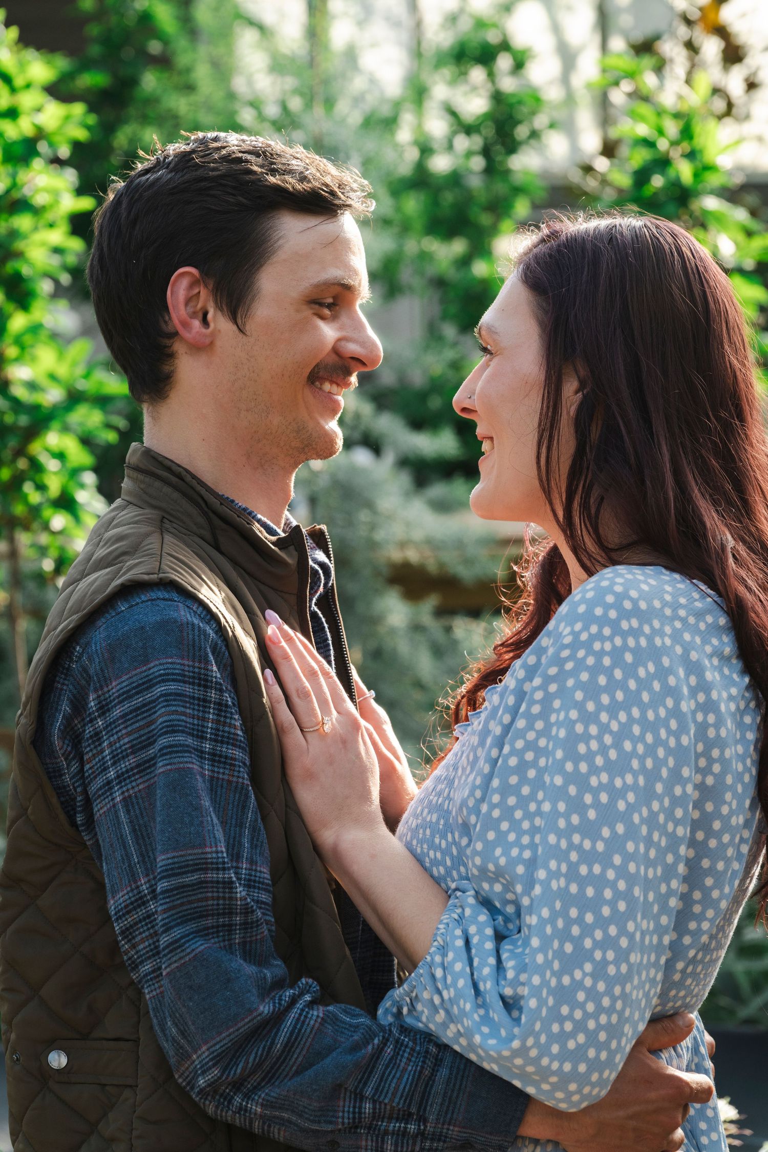 Smiling couple gazing at each other affectionately in a sunlit garden with green foliage.
