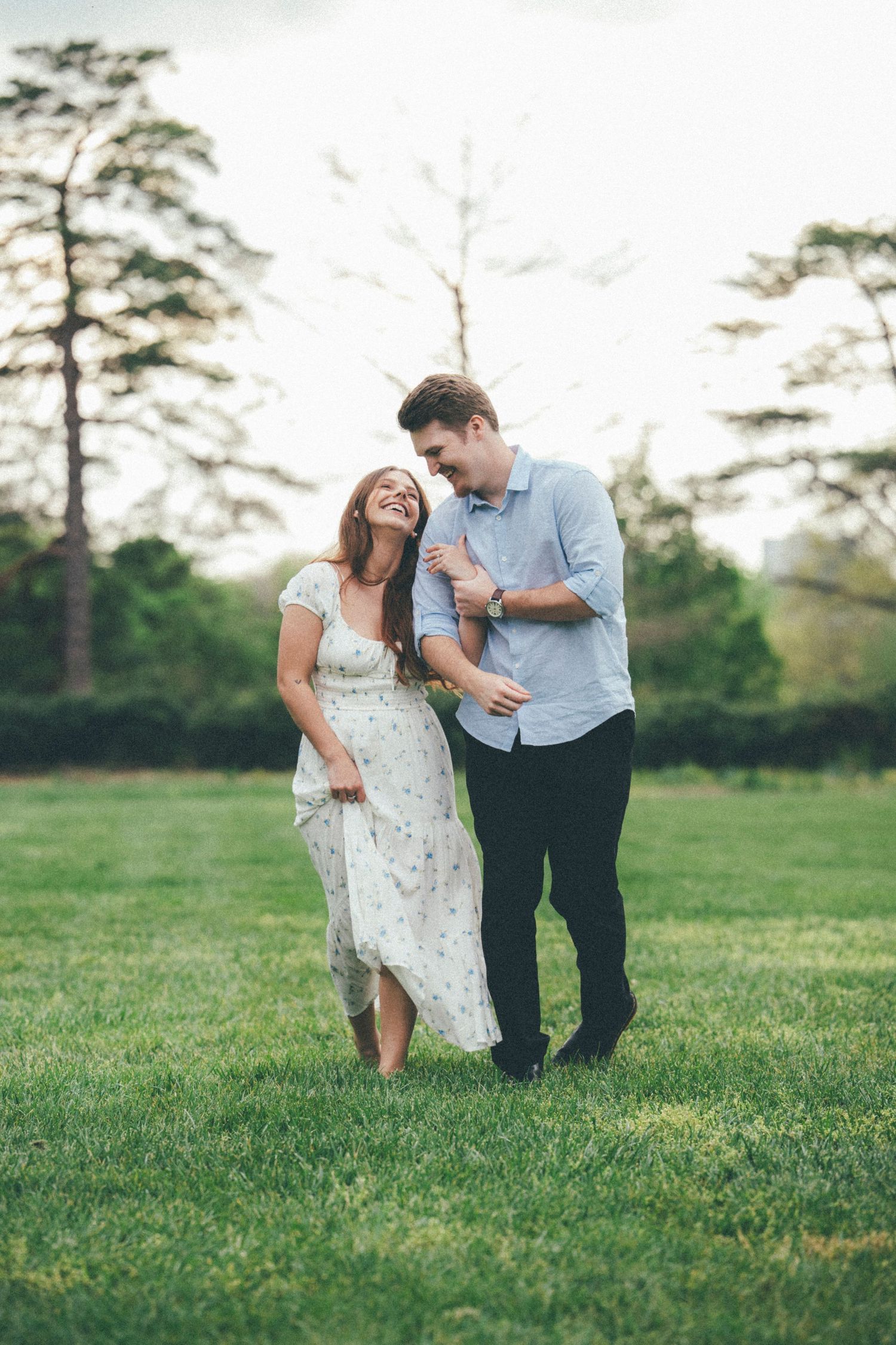 Happy couple laughing and walking together on a lush green lawn surrounded by trees.