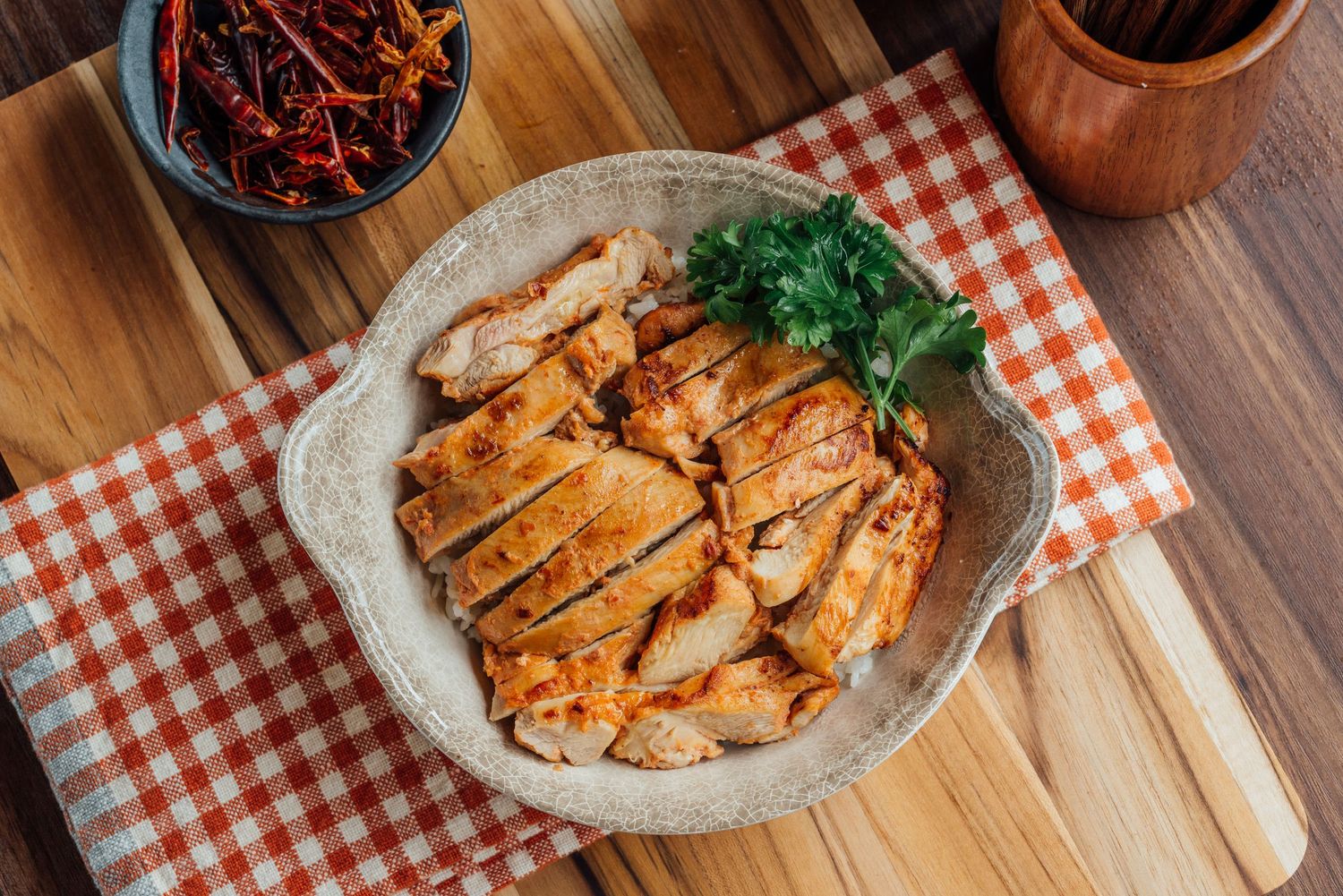 Overhead view of sliced seasoned chicken garnished with parsley in a ceramic bowl on a checkered cloth.