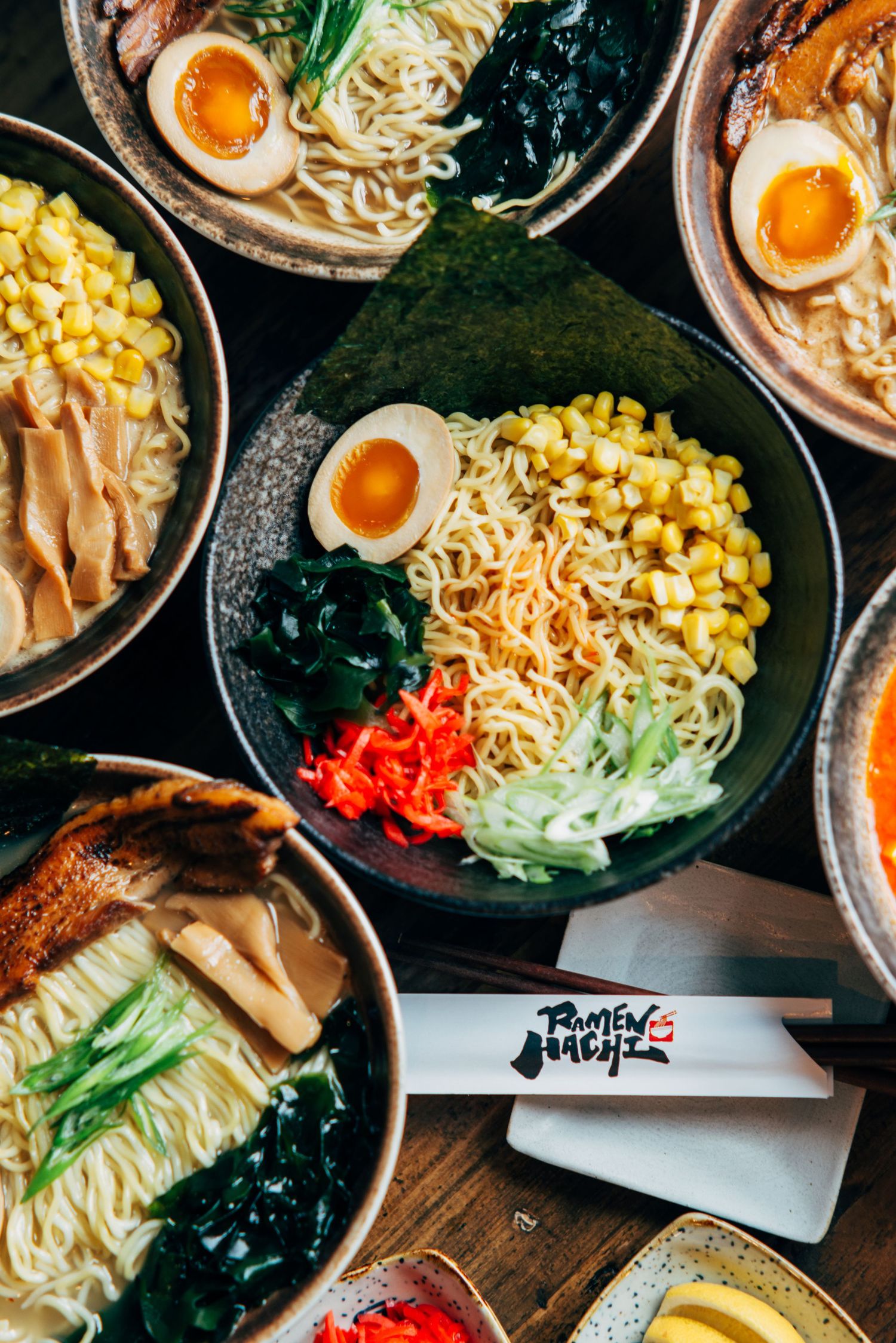 Overhead view of multiple ramen bowls topped with eggs, corn, noodles, and vegetables on a dark table.