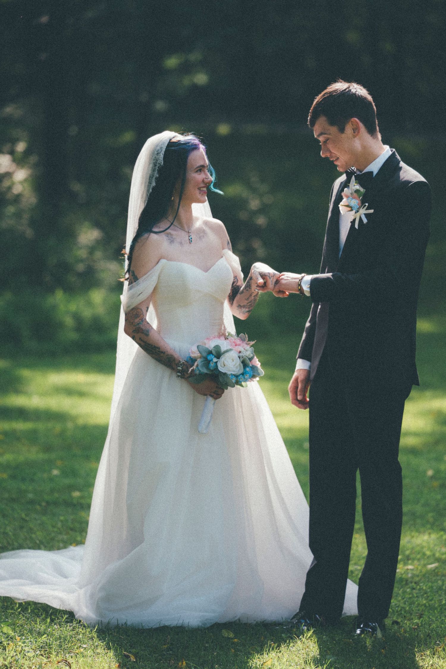 A groom places a wedding ring on his bride's finger outdoors, as she smiles holding a pink and blue bouquet.