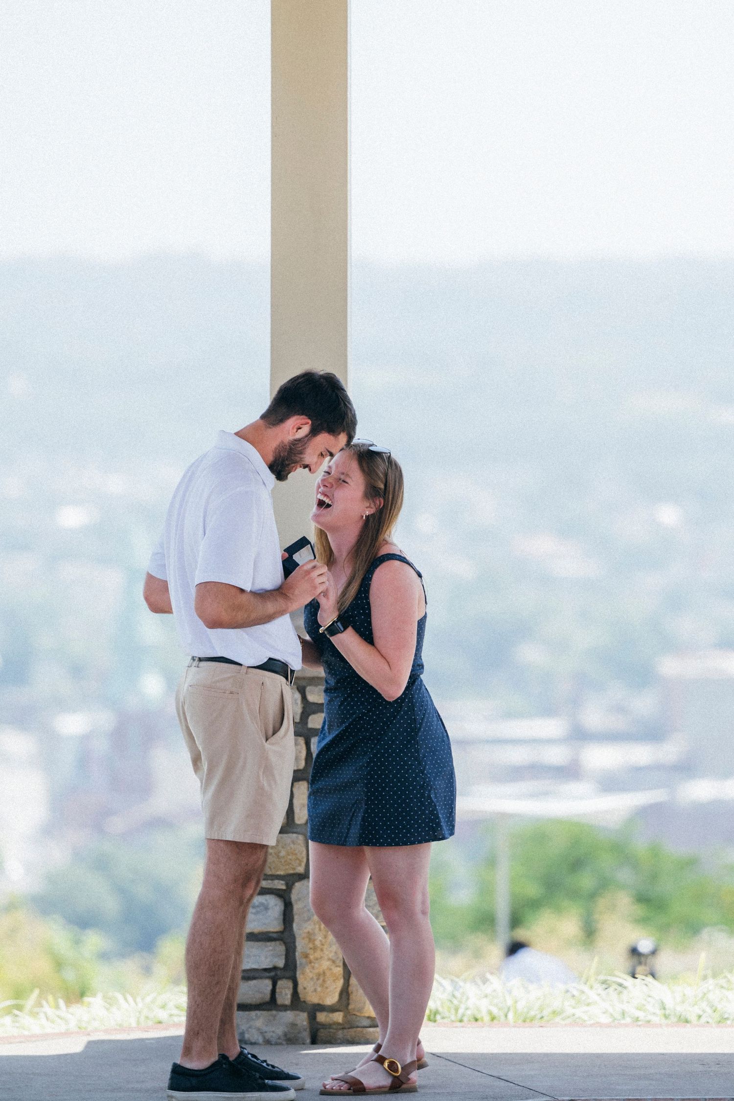 Couple sharing a joyful moment at an outdoor overlook, woman laughing as man holds a small box.