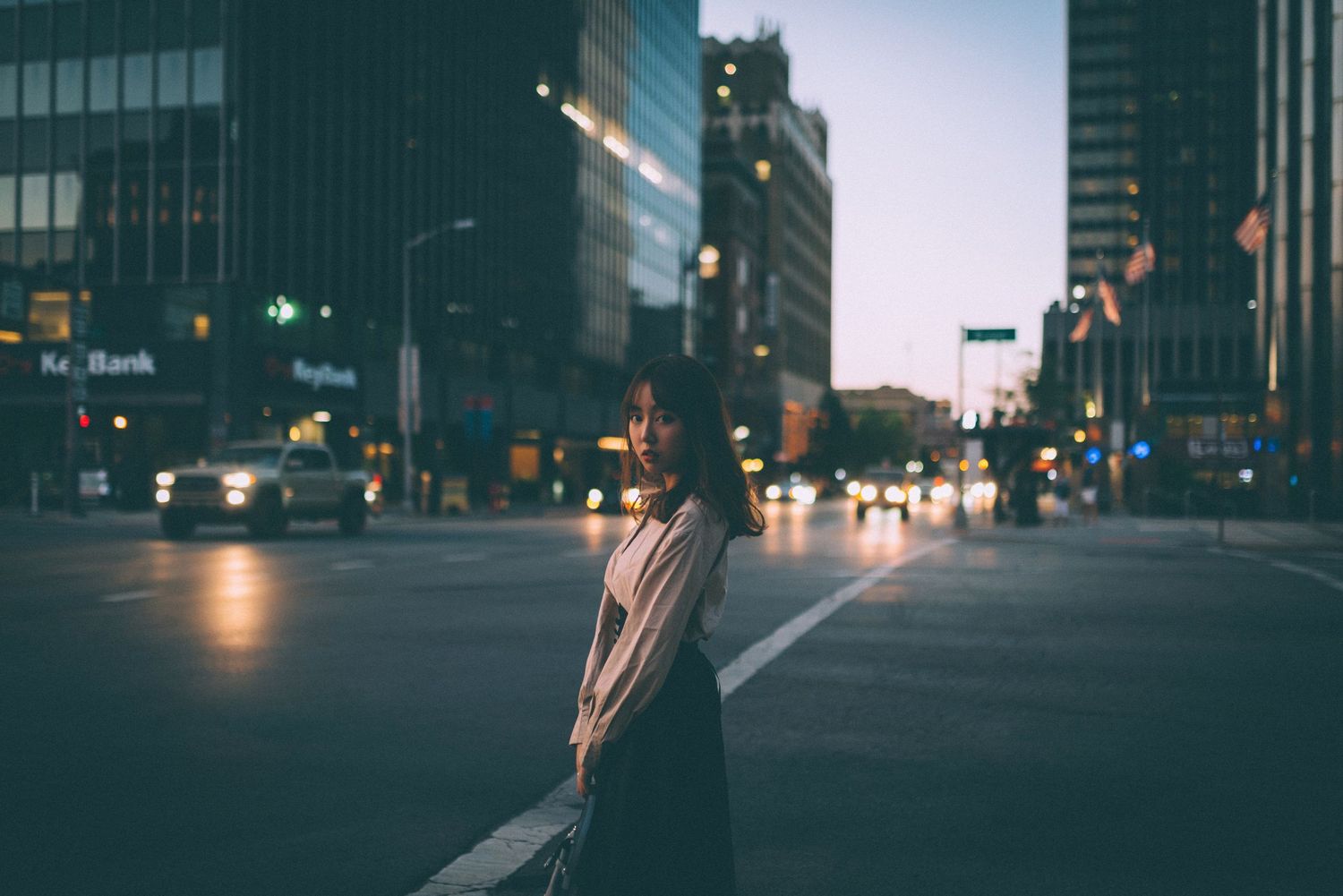 A young woman stands alone on a city street intersection at dusk, with blurred traffic lights and buildings in the background.