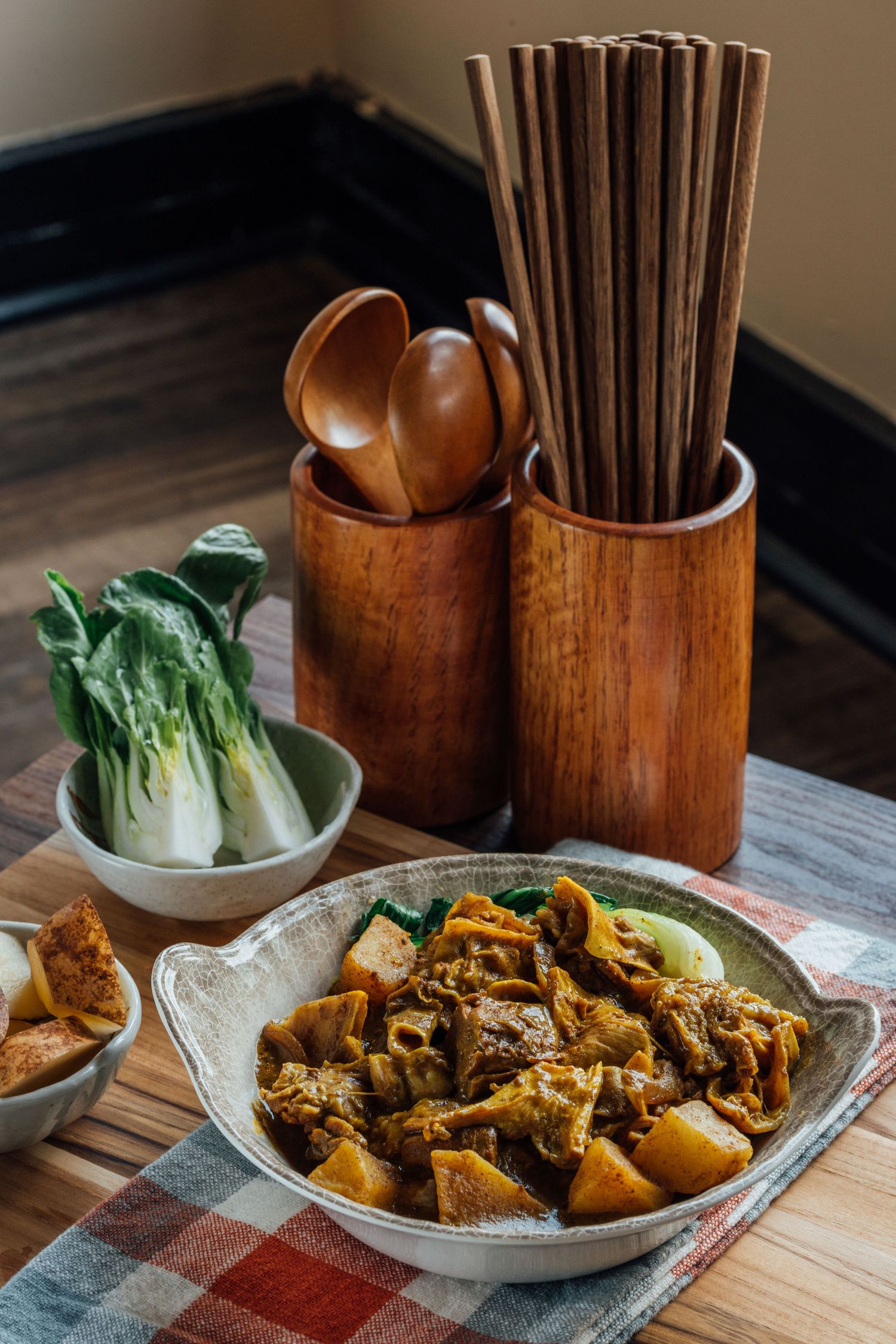 Bowl of curry braised meat and potatoes served with fresh bok choy beside wooden chopsticks and spoons on a table.