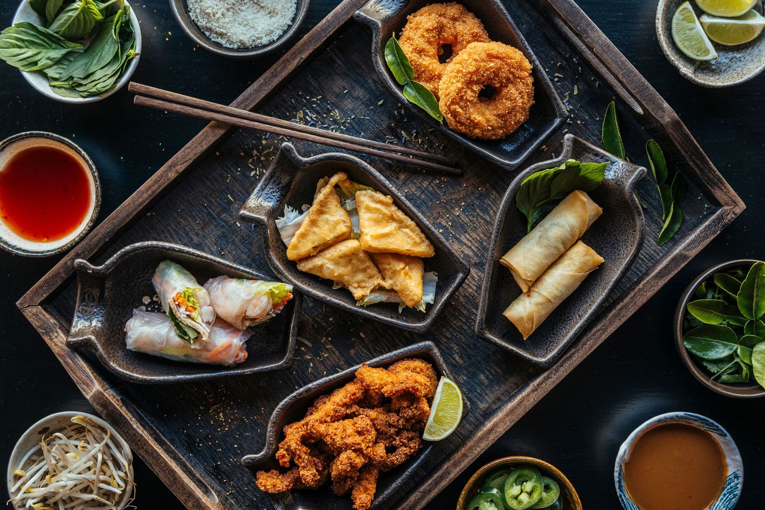 Overhead view of assorted Vietnamese appetizers including spring rolls, fried snacks, and dipping sauces on a rustic wooden tray.