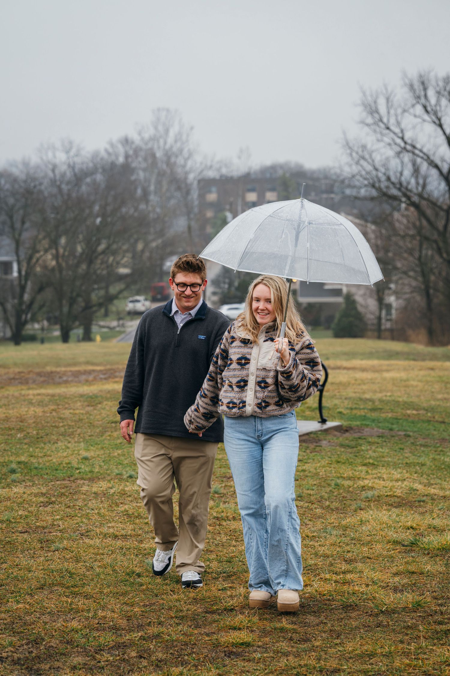 Happy couple walking hand in hand under a clear umbrella on a rainy day in a green park.