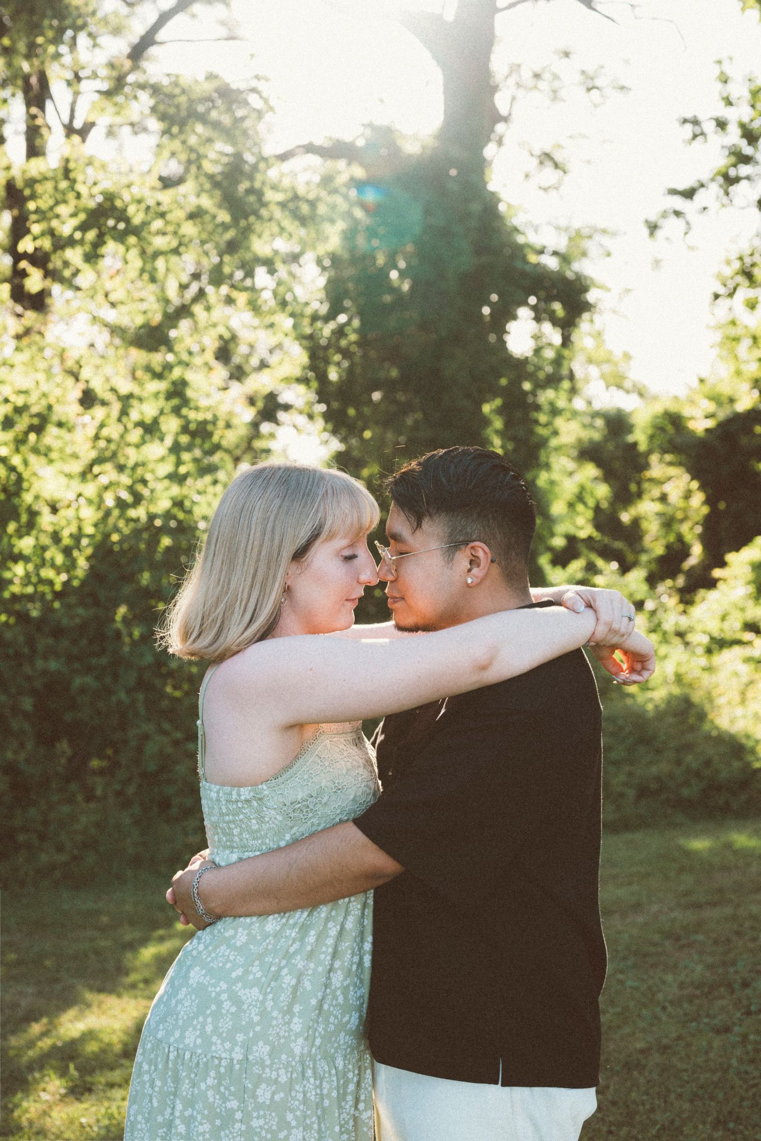 Couple embracing outdoors in golden sunlight, woman in floral dress with arms around partner in black shirt.