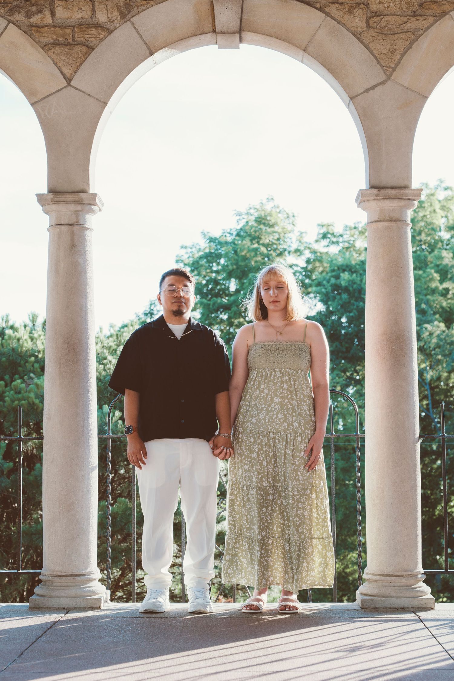 Couple standing hand in hand under stone arches with columns, man in black shirt, woman in floral dress, trees behind.