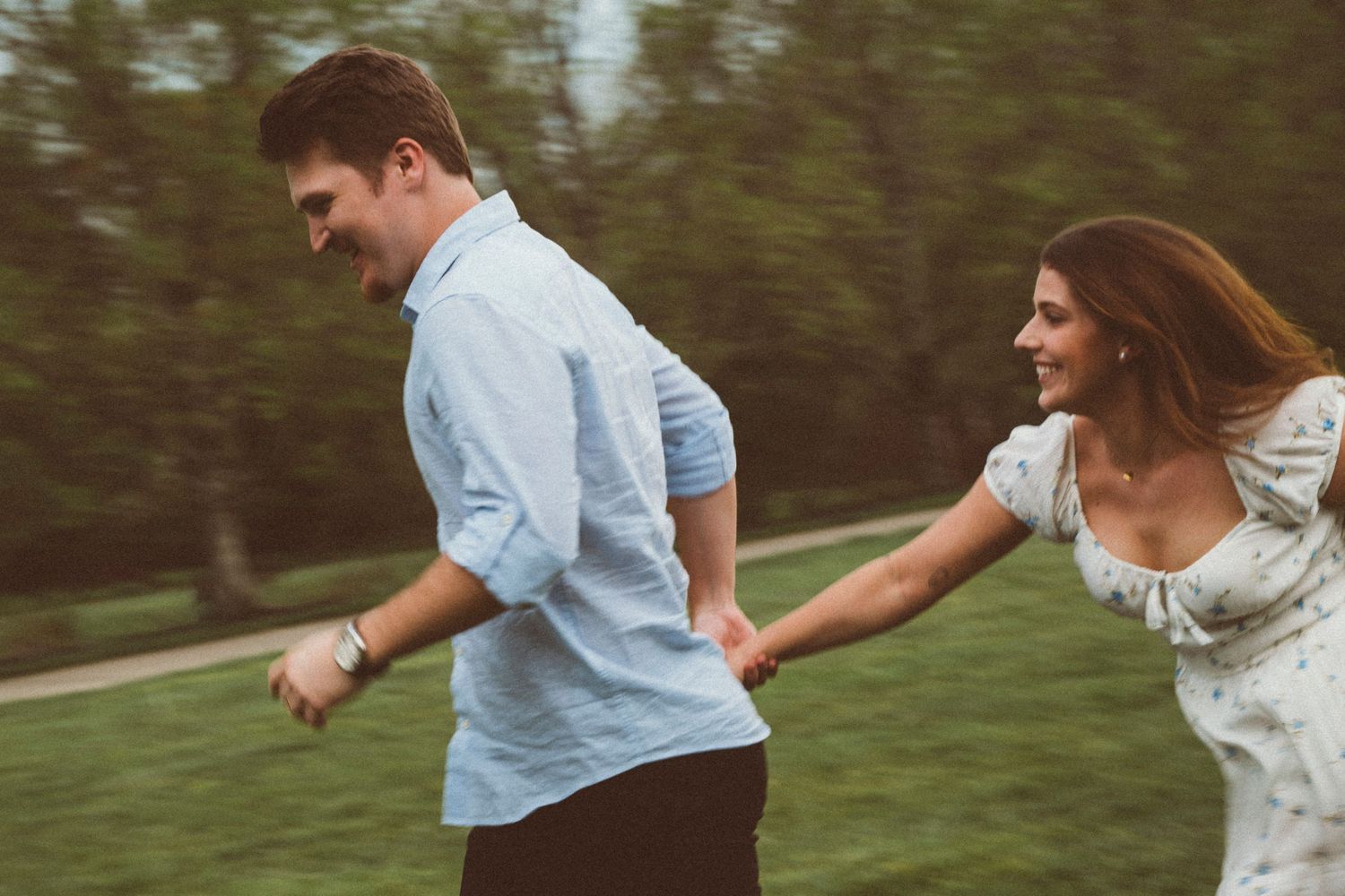 A smiling couple holding hands and running together through a green park on a sunny day.