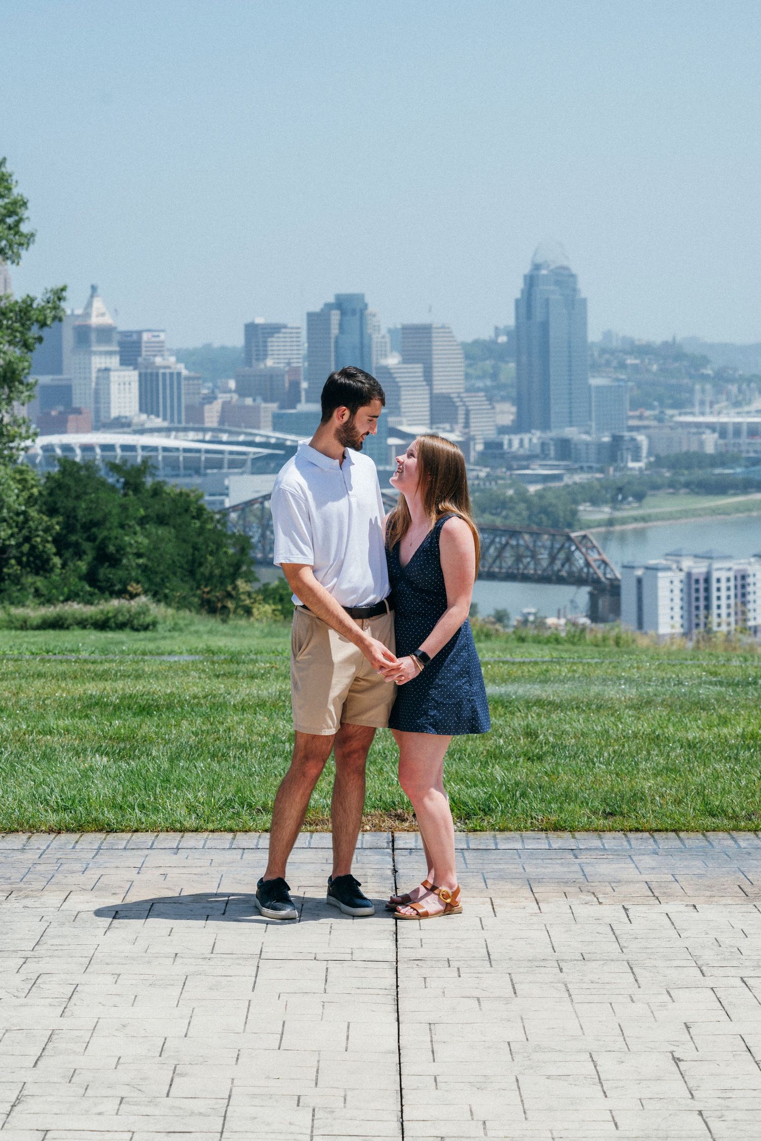 Couple holding hands on a brick path with Cincinnati skyline and Ohio River in the background on a sunny day.