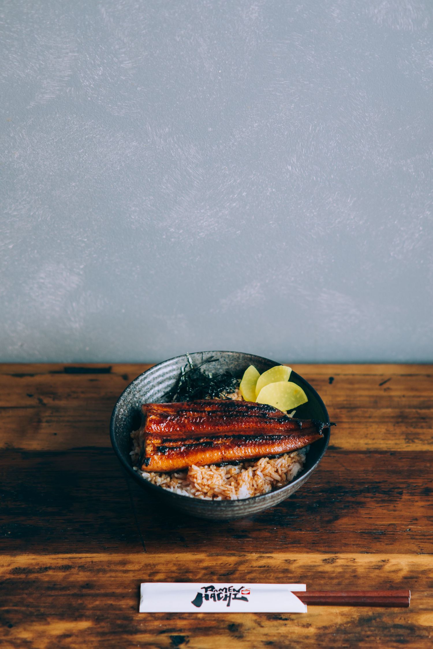 A bowl of unagi eel over rice with lime wedges and chopsticks on a rustic wooden table.