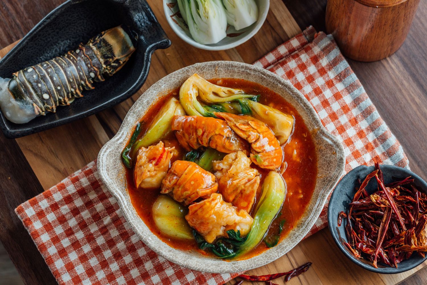 Overhead view of a spicy Korean braised fish dish with bok choy in a clay pot on a checkered tablecloth.