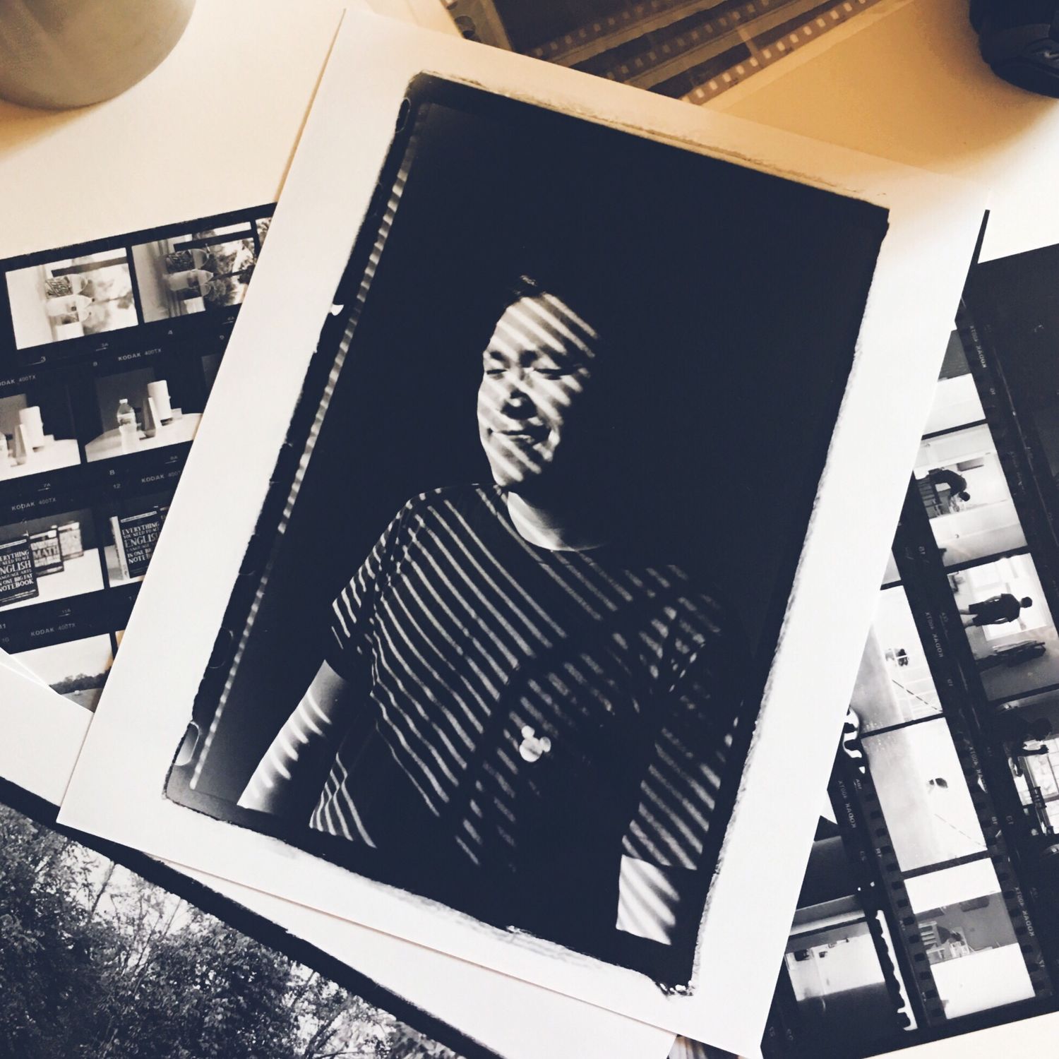 Black and white film photo print of a person with dramatic shadow patterns, surrounded by contact sheets on a desk.