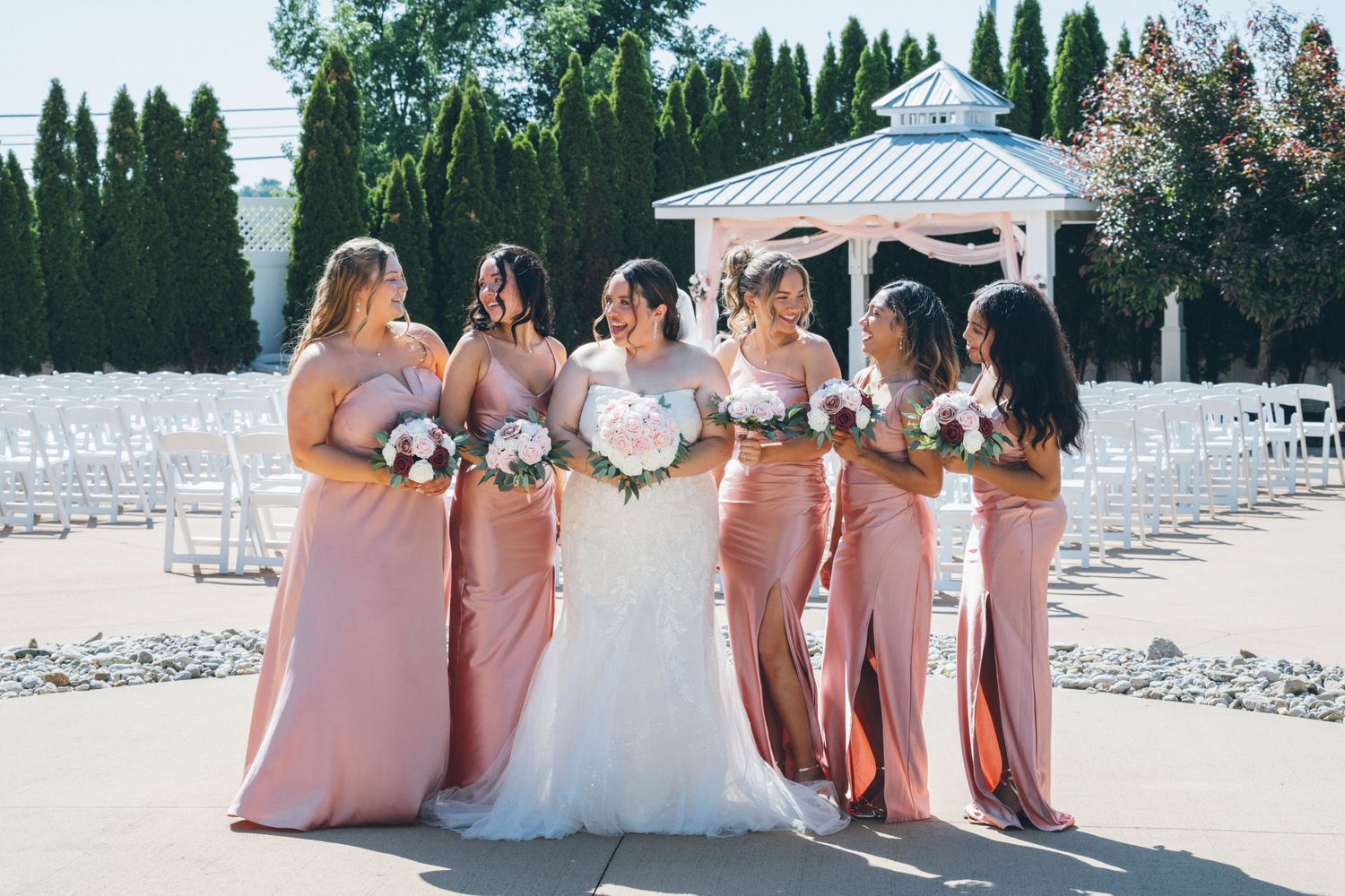 Bride surrounded by bridesmaids in dusty rose gowns holding bouquets near outdoor wedding gazebo.