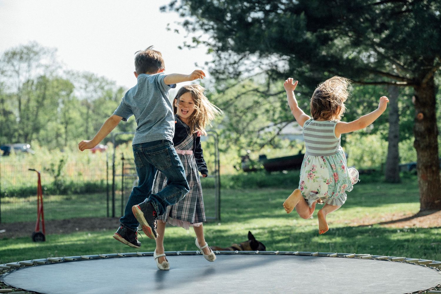 Three children joyfully jumping on a trampoline in a lush green backyard on a sunny day.