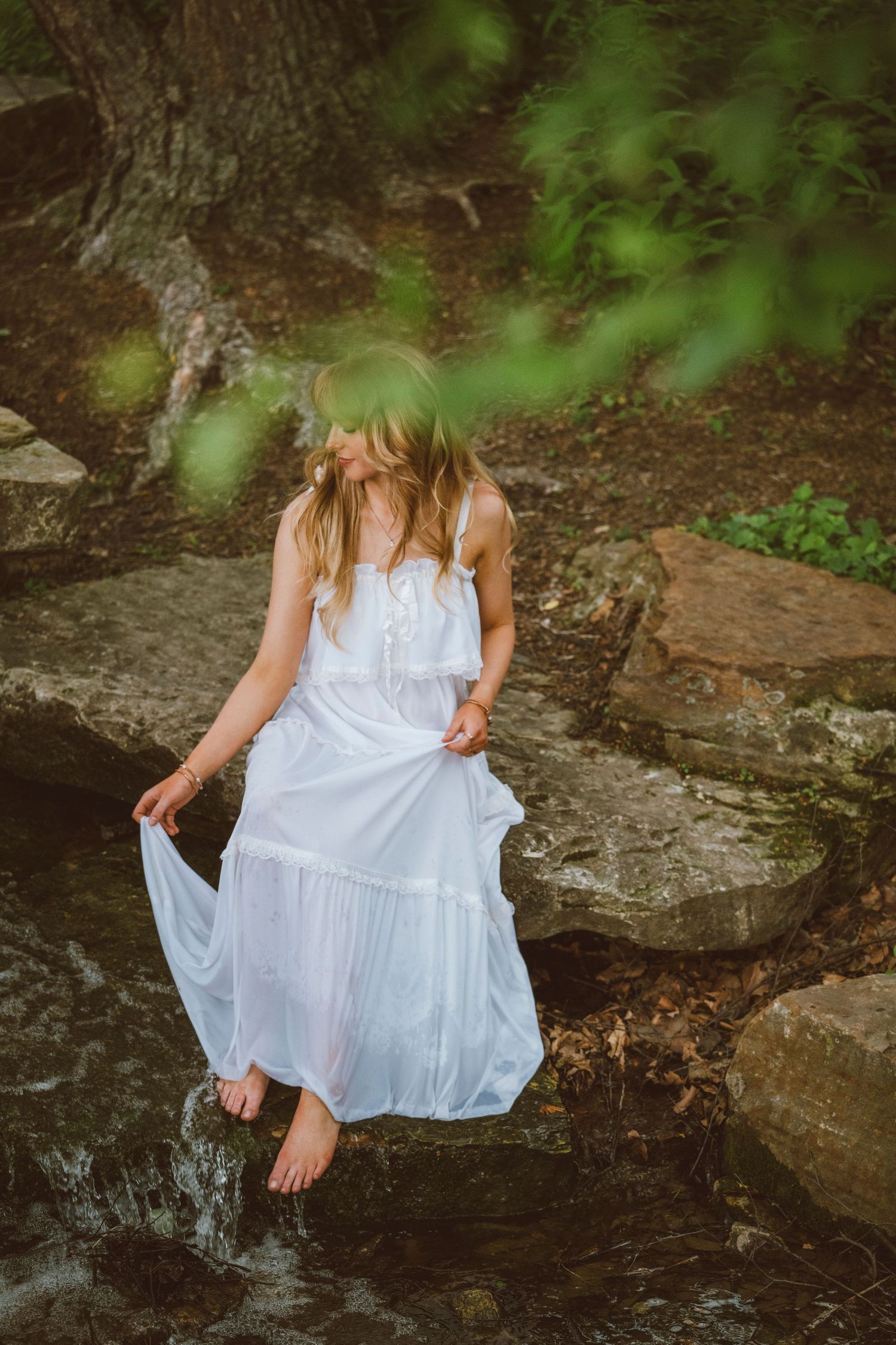 Woman in white dress standing barefoot in a forest stream surrounded by rocks and lush green trees.