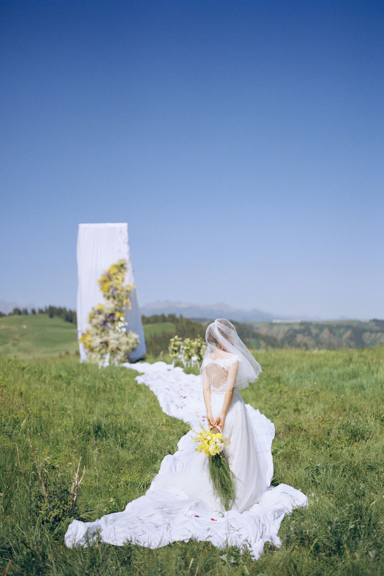 Bride in white gown and veil holds yellow bouquet in green meadow with floral arch under clear blue sky.