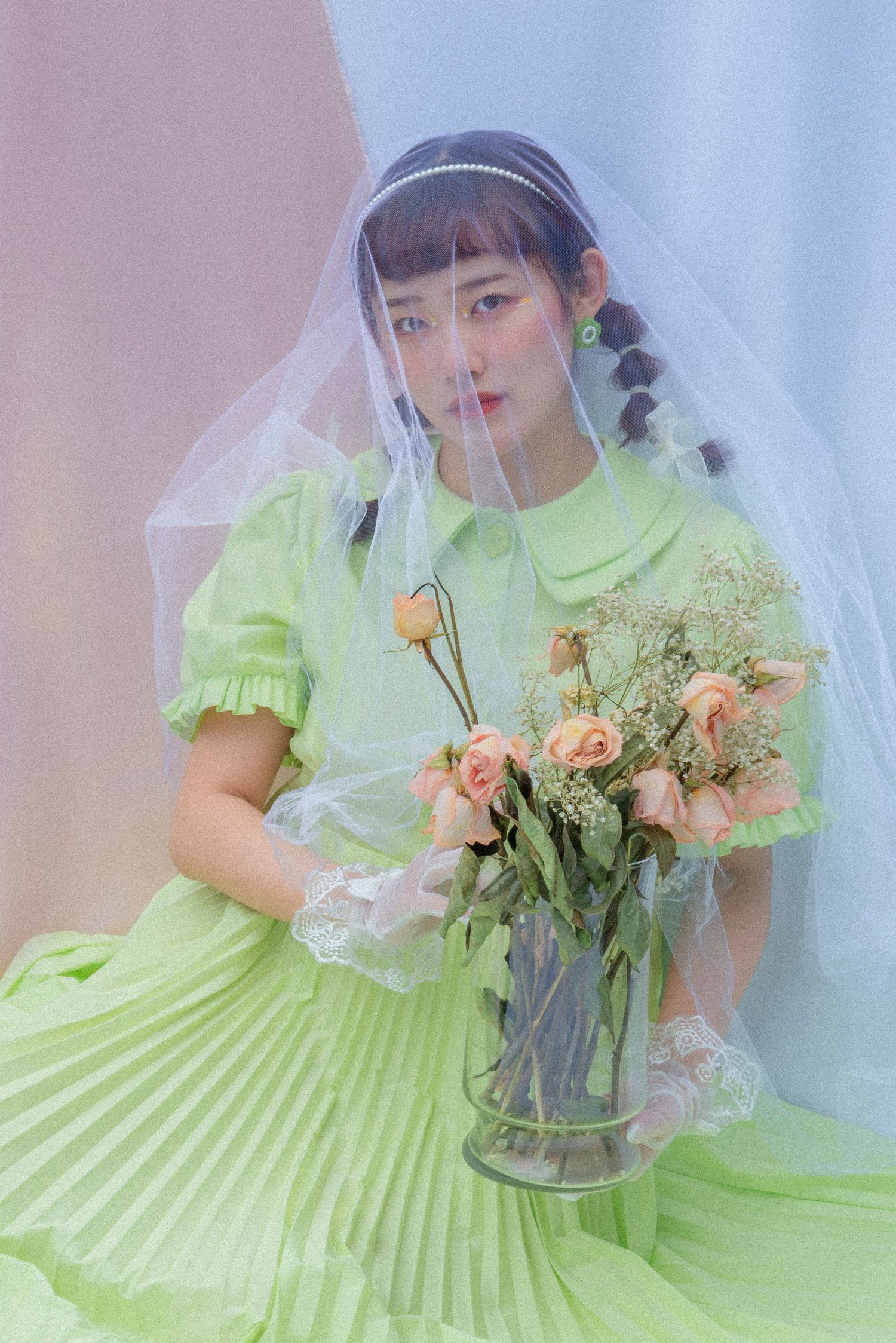Young Asian woman in a lime green dress holds pink roses in a vase while wearing a sheer veil in a pastel studio.