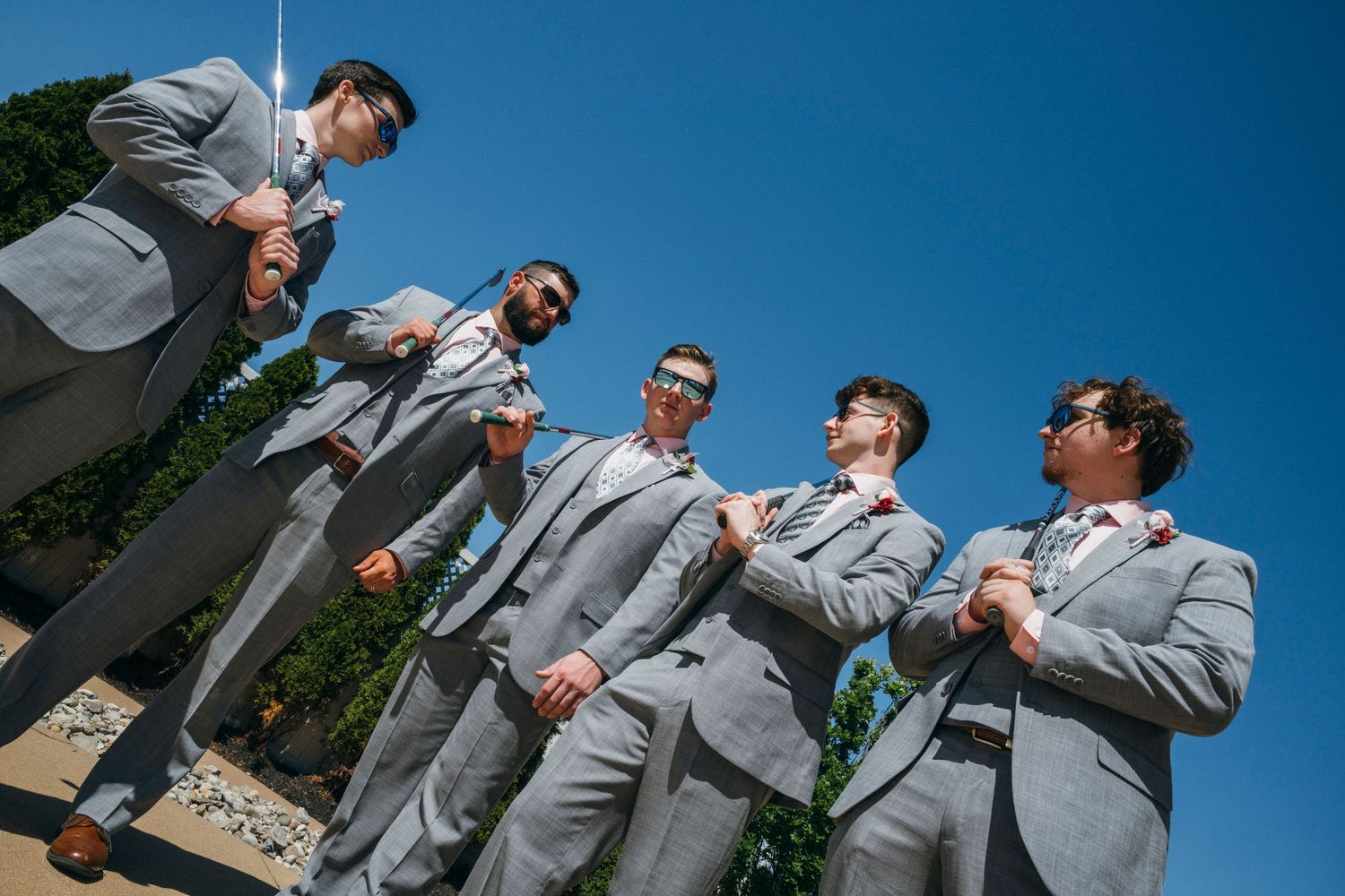 Six groomsmen in matching grey suits and sunglasses posing together outdoors on a sunny wedding day.