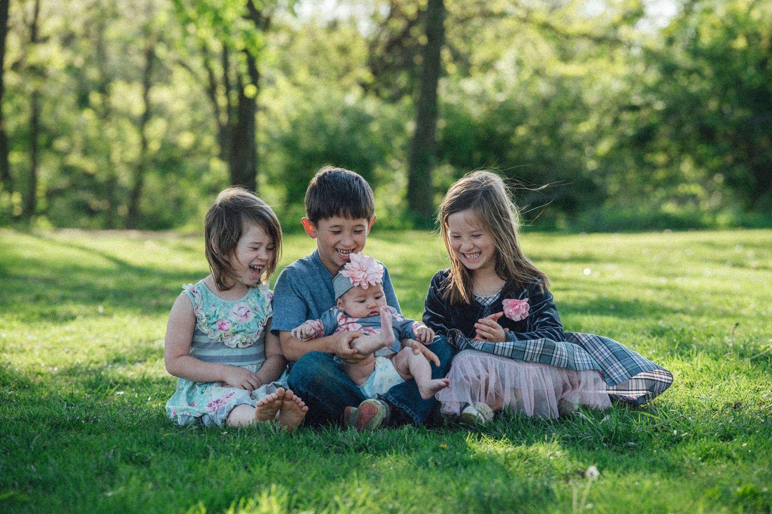 Three happy children sitting on green grass in a sunny park, smiling and holding a baby outdoors.