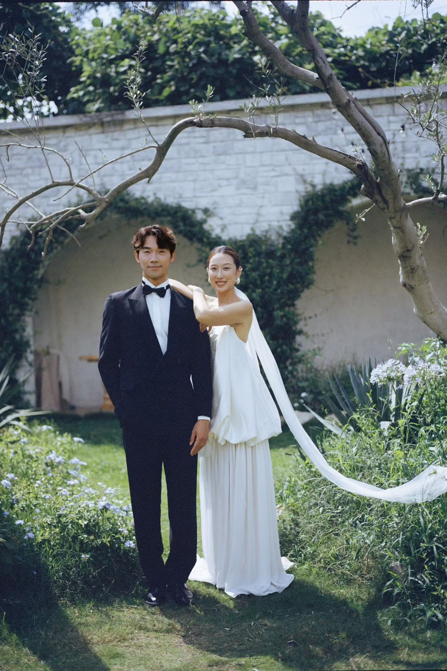 Couple in wedding attire posing in lush garden with groom in black tuxedo and bride in white gown.