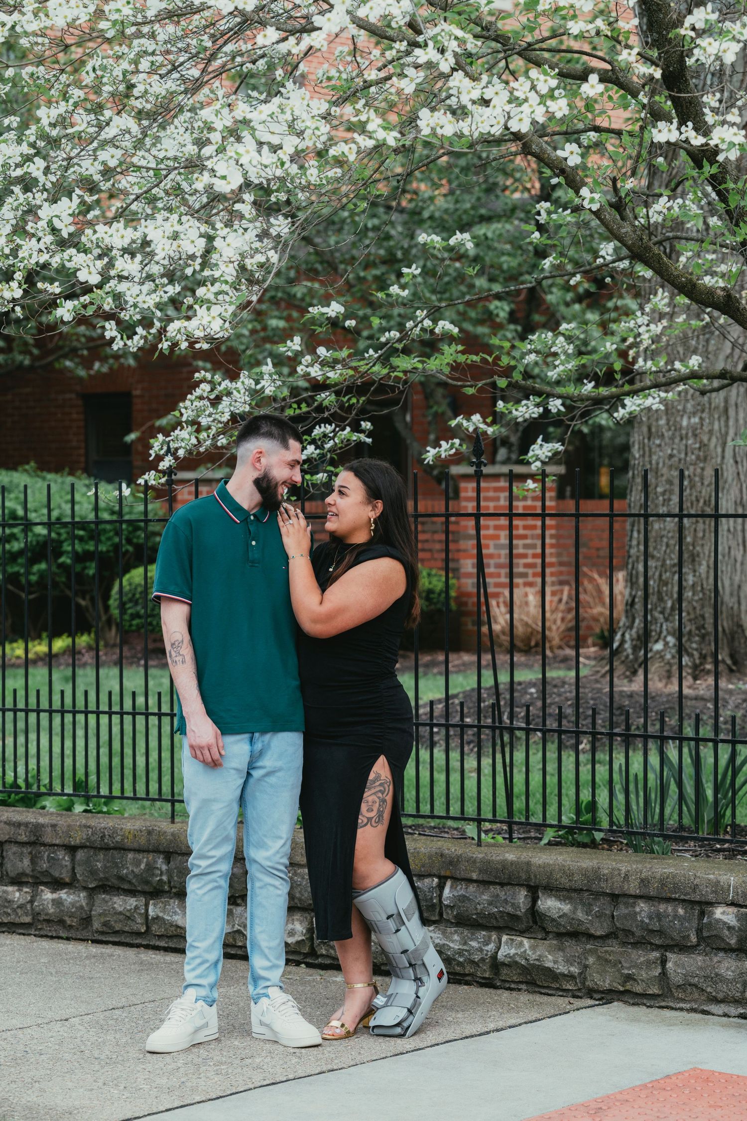 Couple smiling at each other under blooming white cherry blossom tree by iron fence.