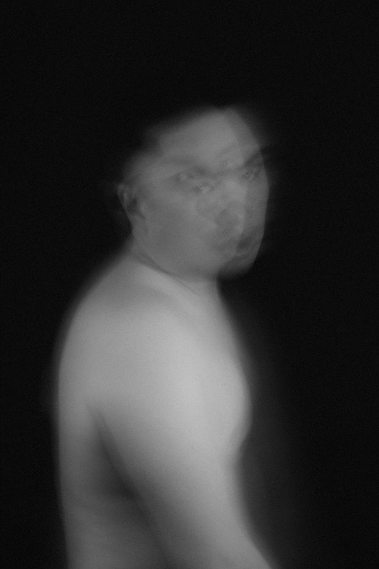 Blurry black and white portrait of a shirtless person wearing glasses against a dark background.