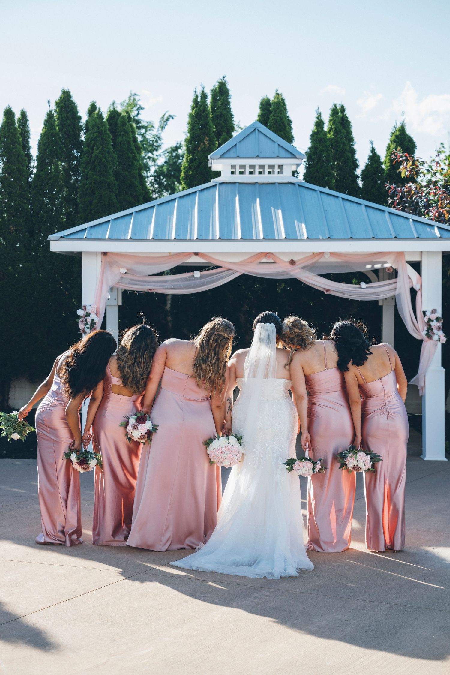 Bride with bridesmaids in blush pink gowns standing together facing elegant white wedding gazebo.