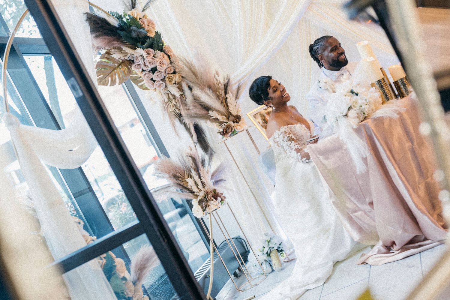 Elegant wedding couple in white attire standing at decorated altar with floral arrangements and draped fabric.