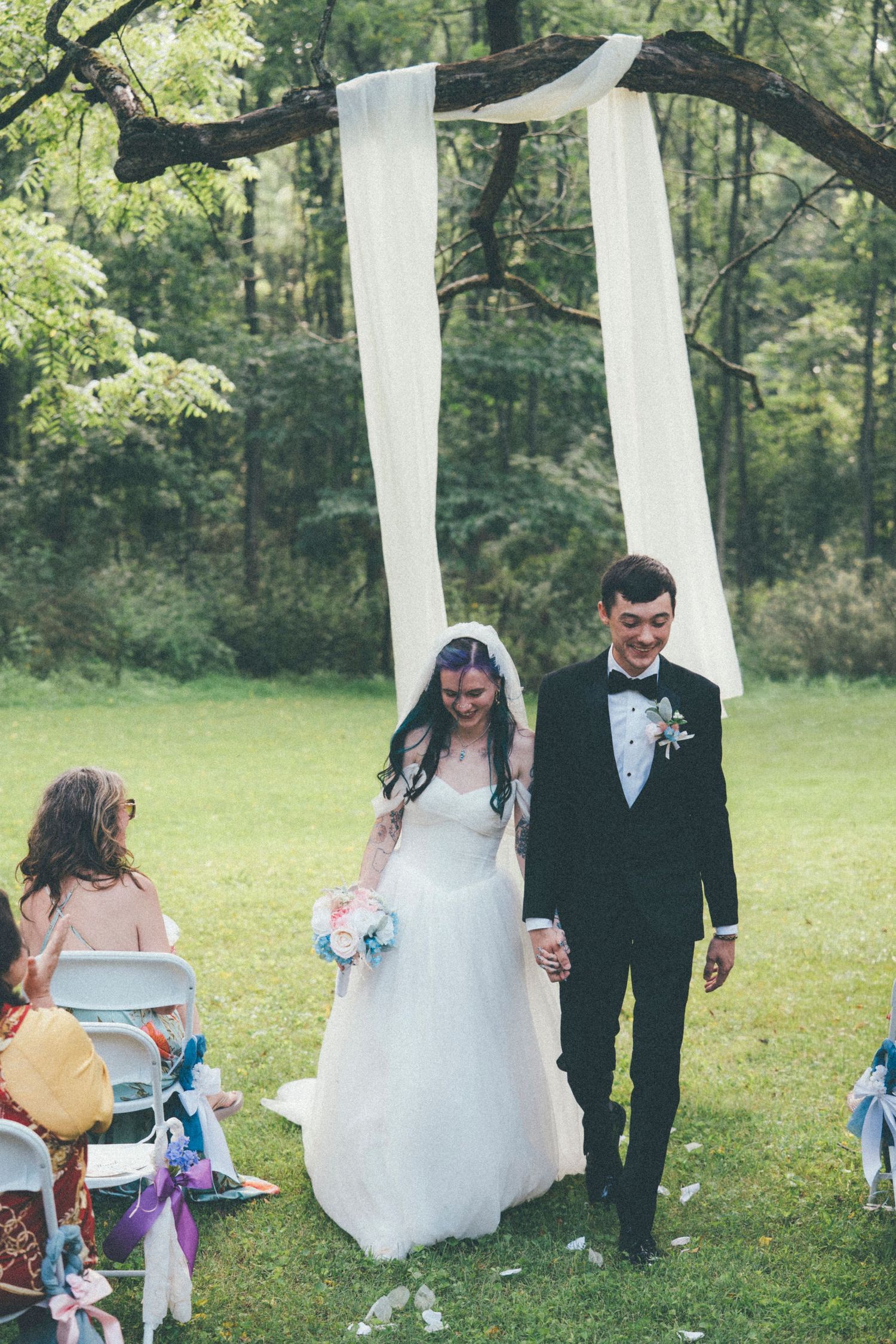 Bride and groom walk hand-in-hand down outdoor aisle after wedding ceremony beneath draped white fabric arch.
