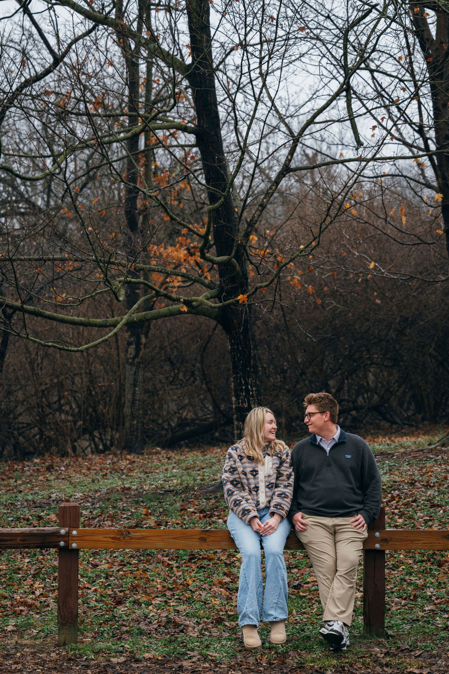 Couple sitting on a wooden fence in an autumn park, sharing a warm smile among bare trees.