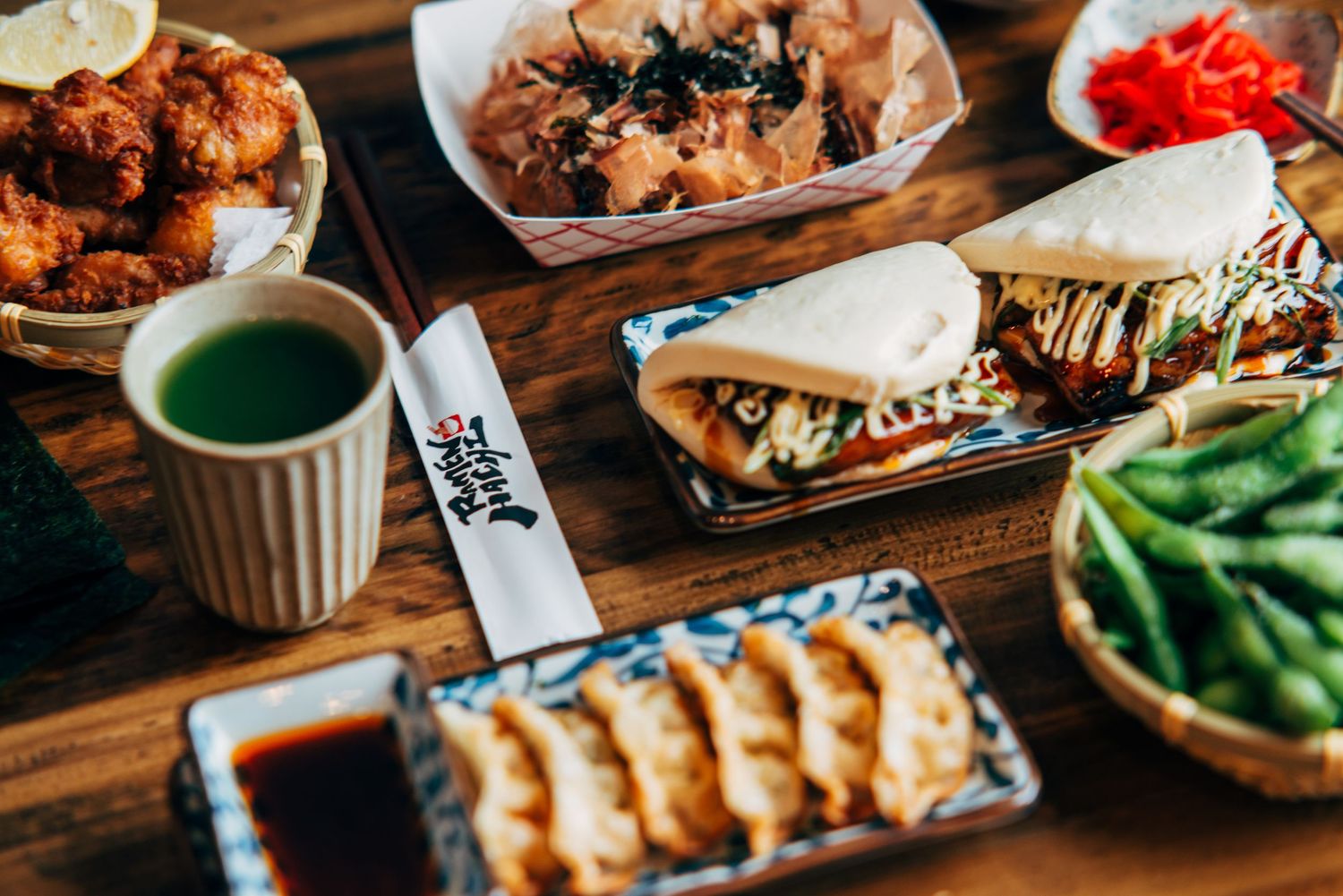 A spread of Japanese dishes including gyoza, bao buns, fried chicken, and green tea on a wooden table.