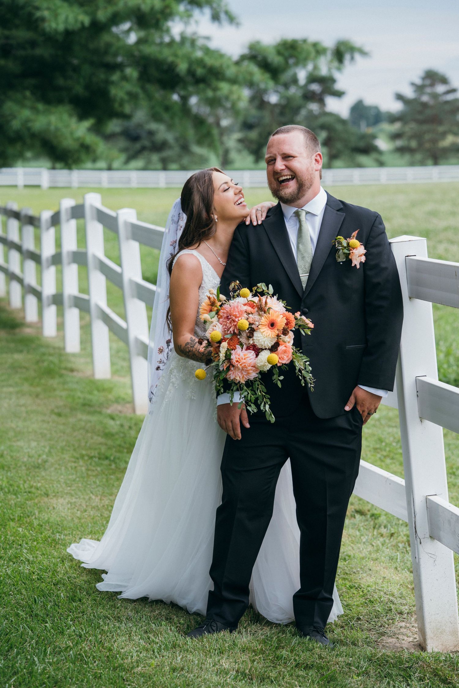 Bride and groom laughing together by white fence on green farm, bride holding colorful pink bouquet in wedding dress.