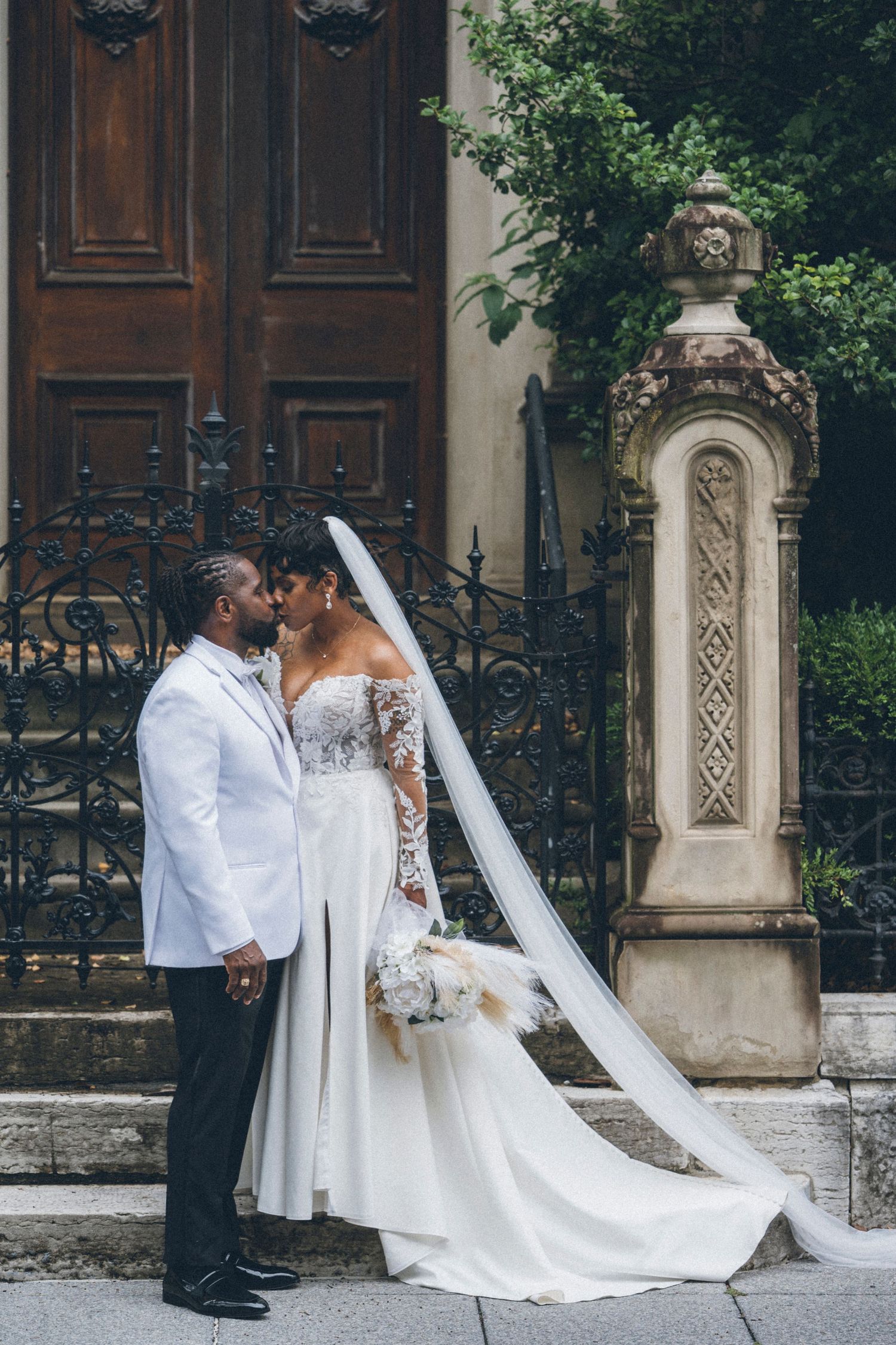 A bride and groom share a romantic kiss outside an ornate building, she in a lace gown with veil, he in a white suit.