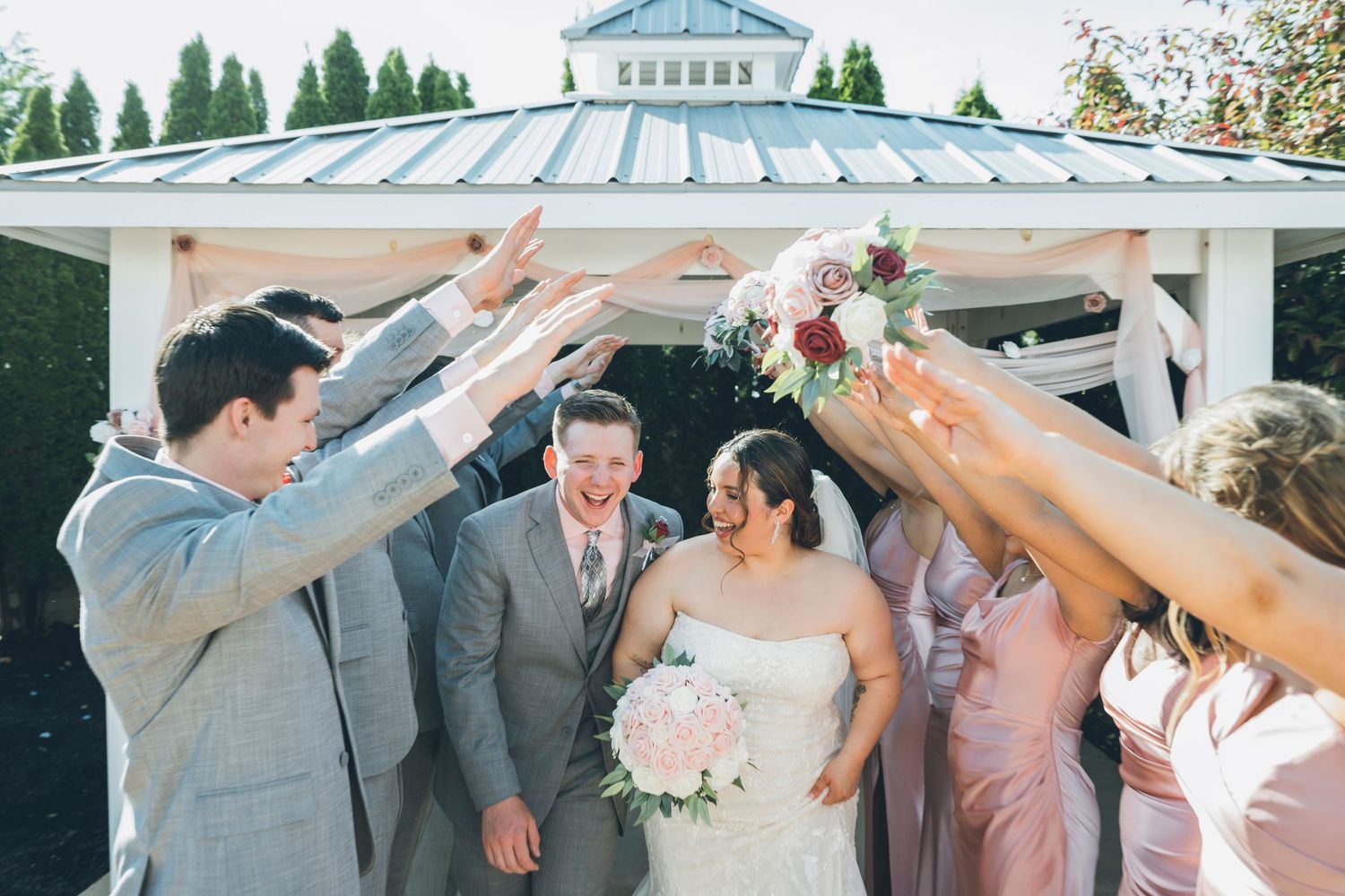Smiling newlyweds celebrated by wedding party raising hands under gazebo on their wedding day.
