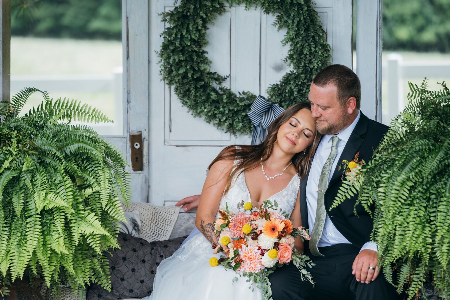 Bride and groom sitting on a porch surrounded by ferns and a wreath, sharing a romantic moment on their wedding day.