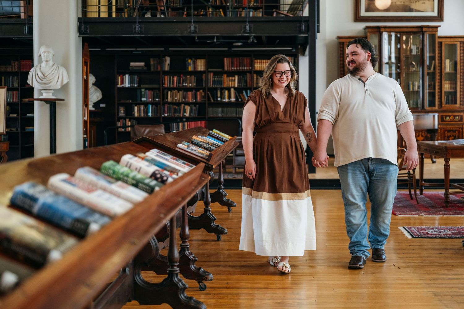 Couple holding hands walking through a vintage library with wooden bookshelves and hardwood floors.