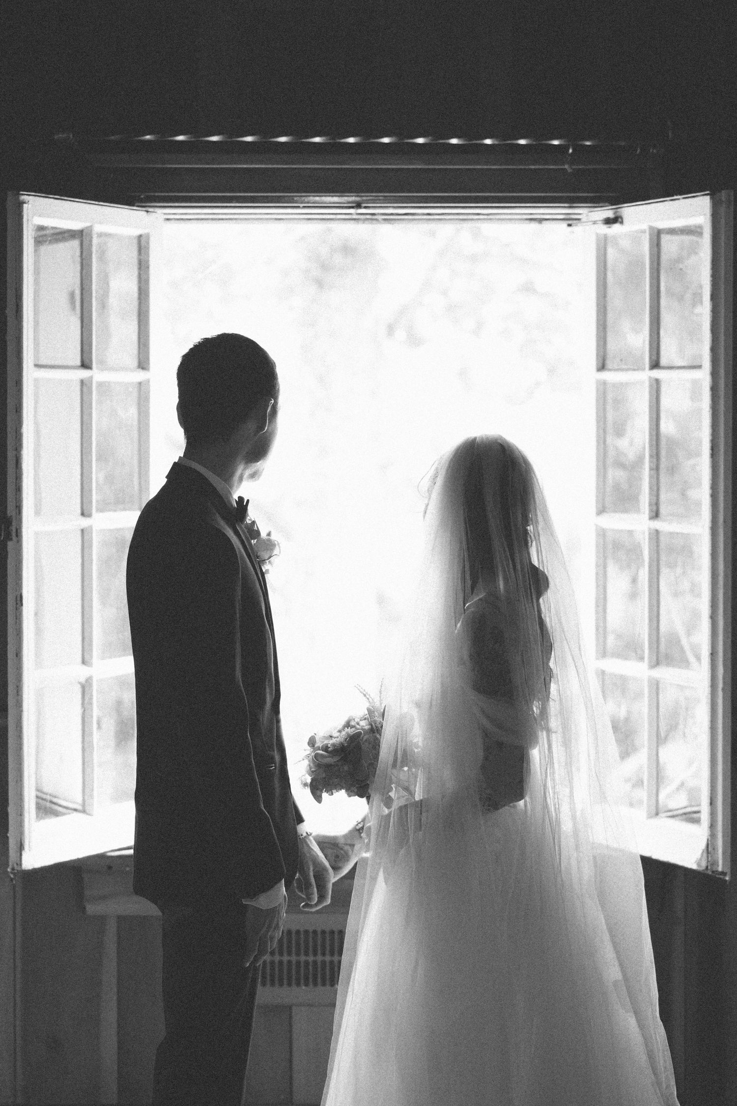 Black and white photo of bride and groom silhouettes holding hands before open French doors on wedding day.