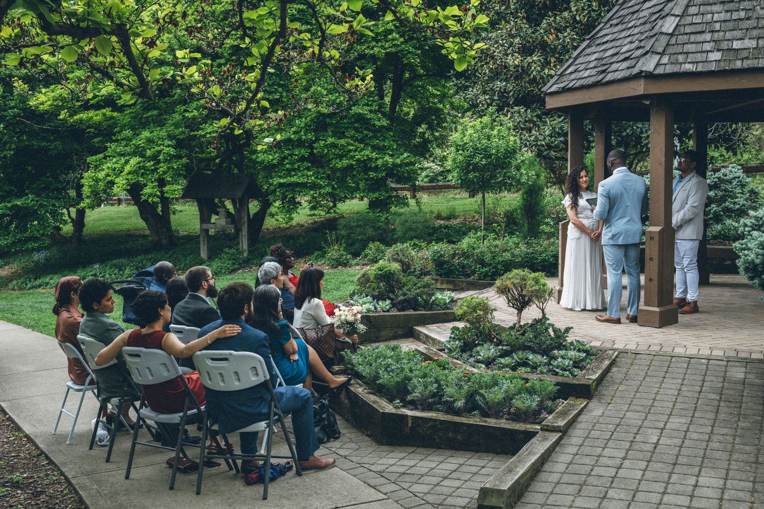 Small intimate outdoor wedding ceremony at garden gazebo with guests seated on folding chairs.