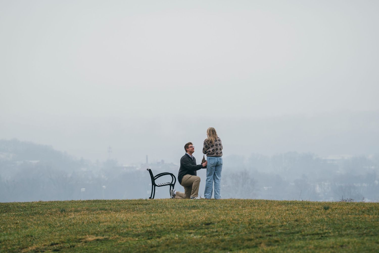 Man proposing on one knee to woman on a hilltop with a misty cityscape behind them.