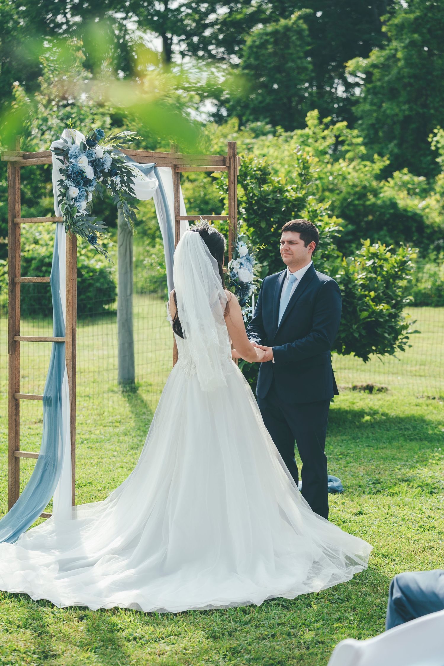 Bride and groom exchange vows outdoors under a wooden arch adorned with blue florals, surrounded by lush greenery.