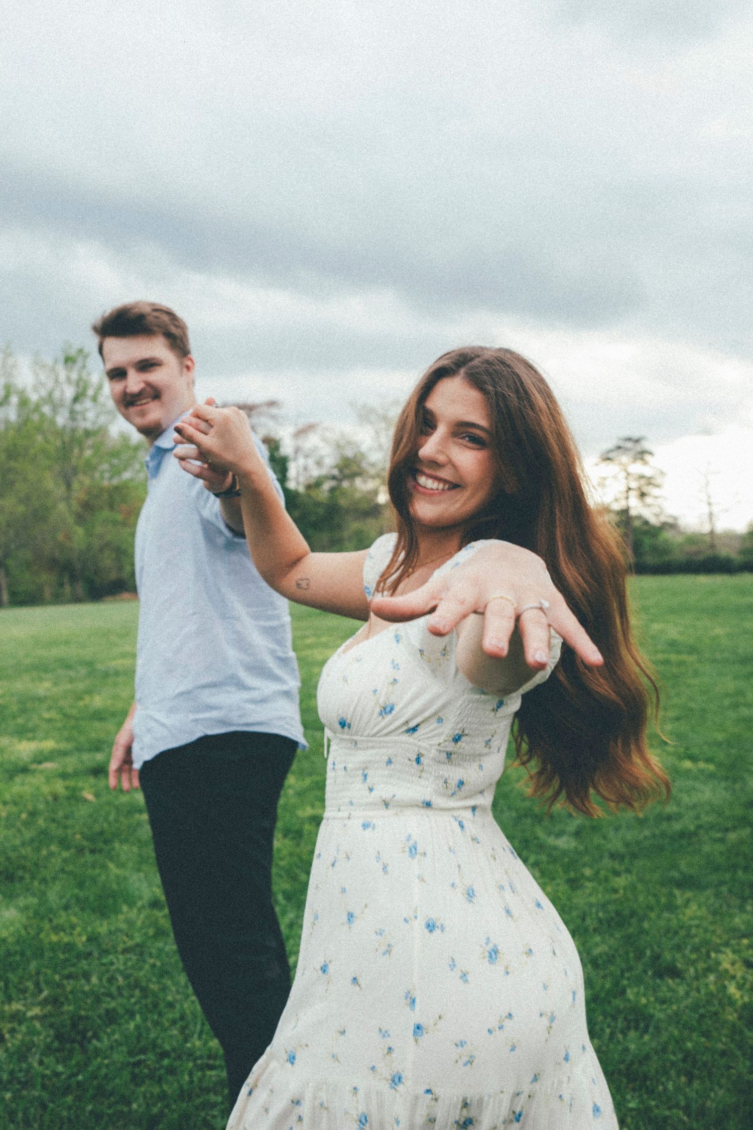 Smiling woman in floral dress shows engagement ring while holding mans hand in green field.