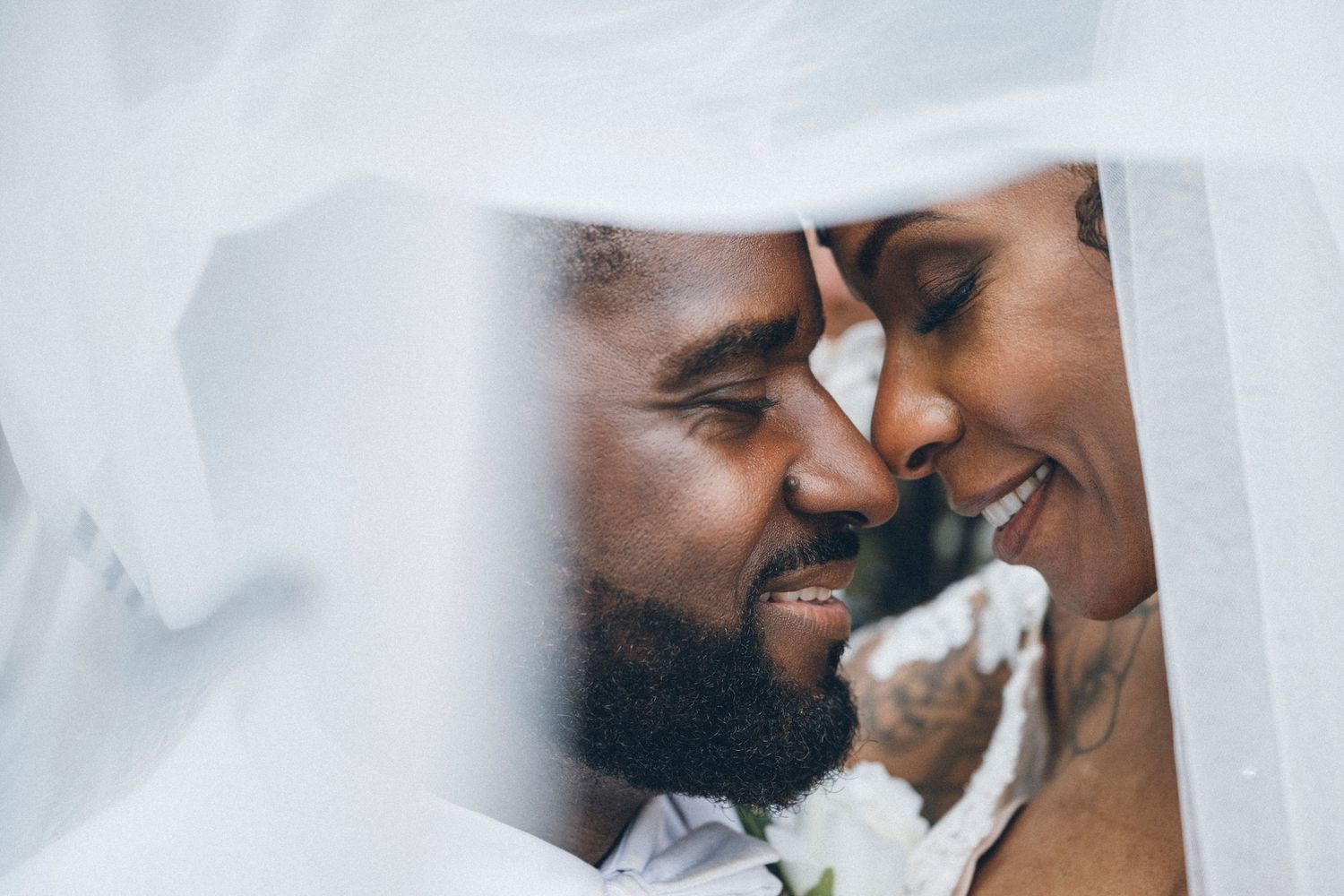 A romantic couple shares an intimate moment under a white wedding veil, foreheads touching and smiling softly.