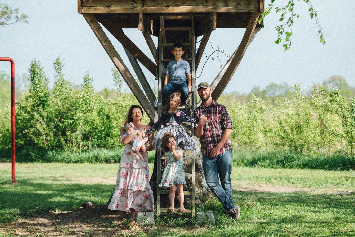 Family of five posing together under a wooden treehouse in a lush green backyard on a sunny day.