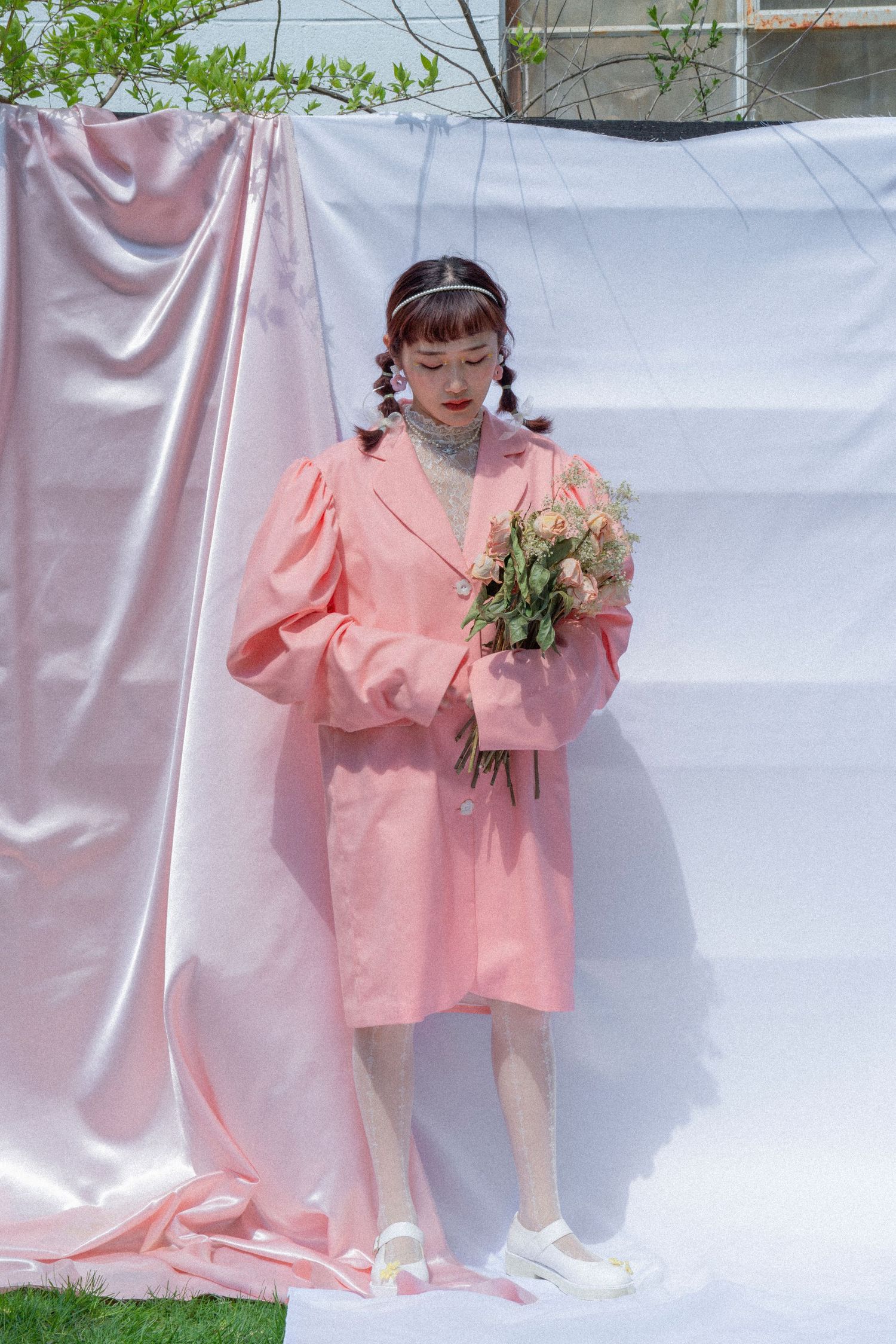 Young Asian woman in a pink oversized blazer dress holds a bouquet of roses against draped pink fabric outdoors.